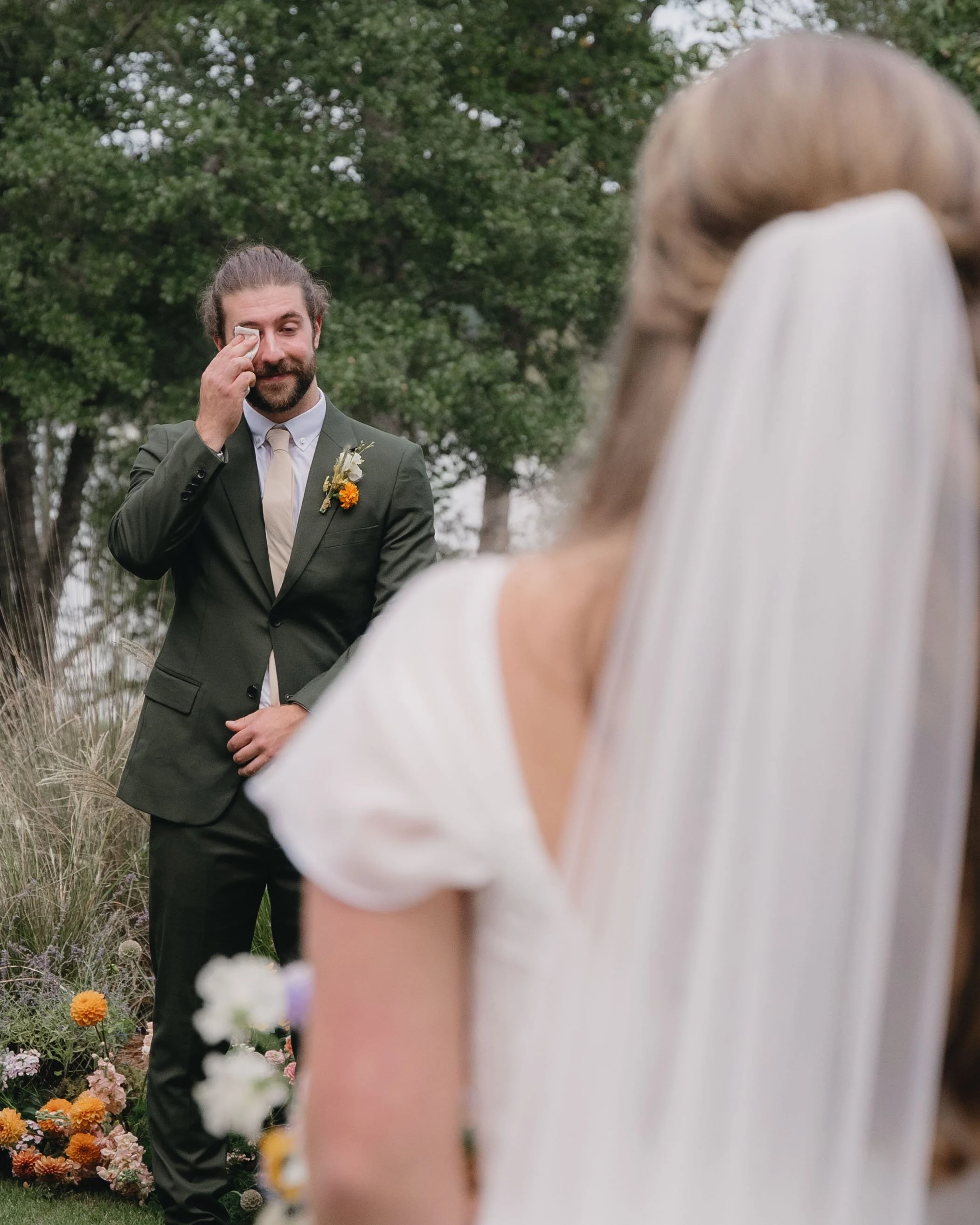A groom wiping a tear during a wedding ceremony outdoors with trees and flowers.