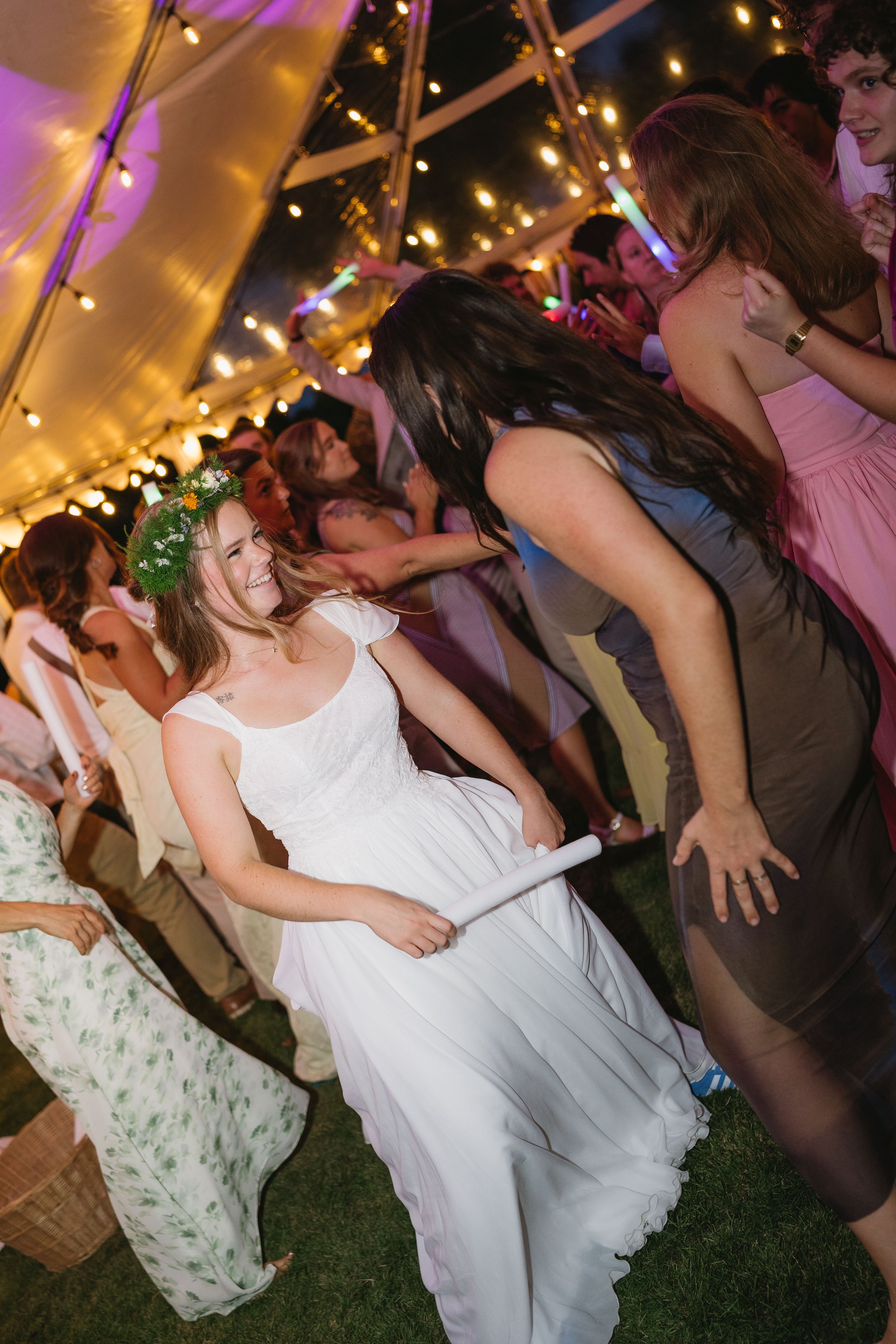 A woman in a white dress with a floral crown is holding a rolled-up paper and smiling at another woman in a black dress, both dancing at a wedding reception inside a lit tent with string lights.