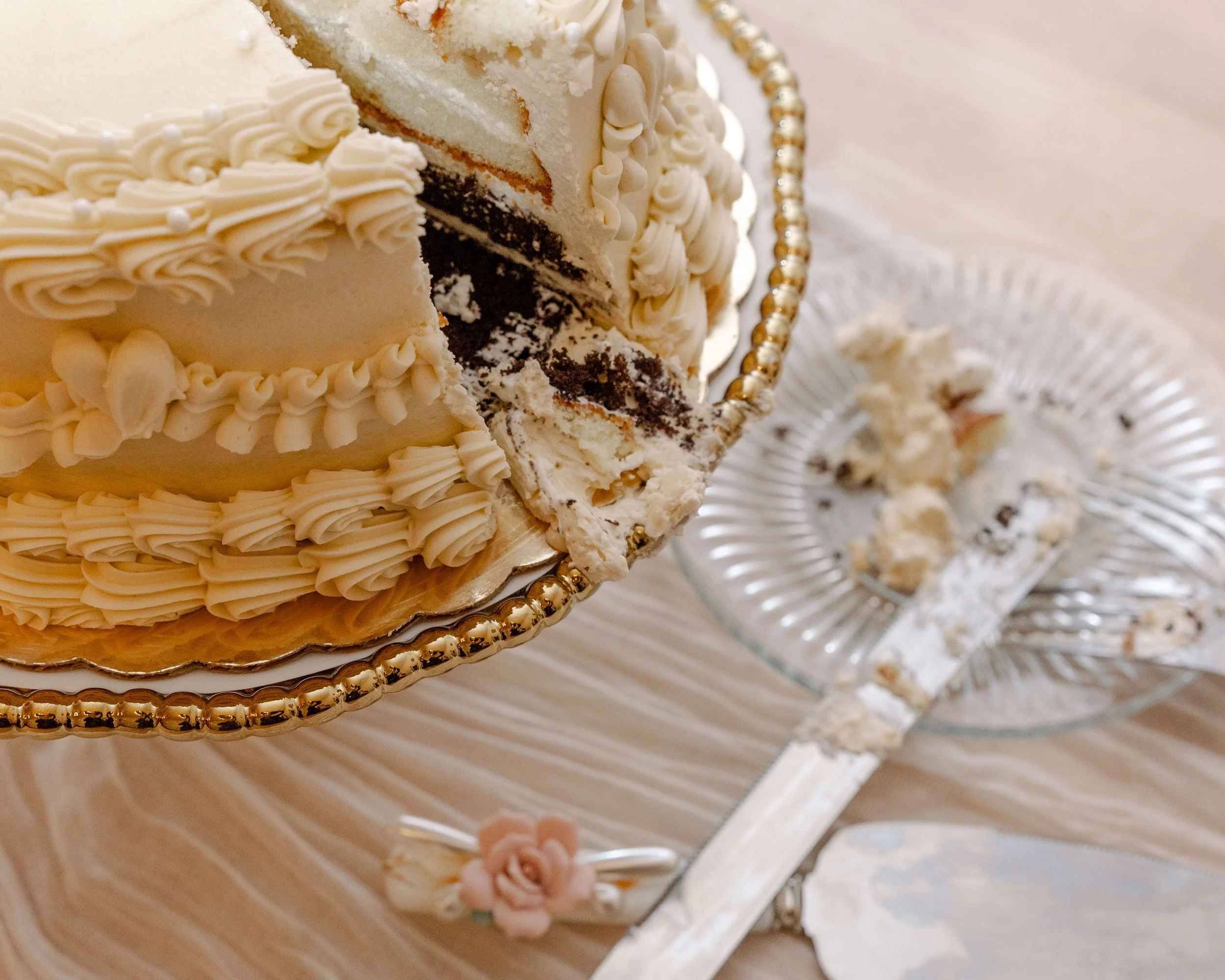 A partially sliced white cake with decorative icing on a gold-edged cake stand, with a glass dish of cake pieces and cake server nearby.