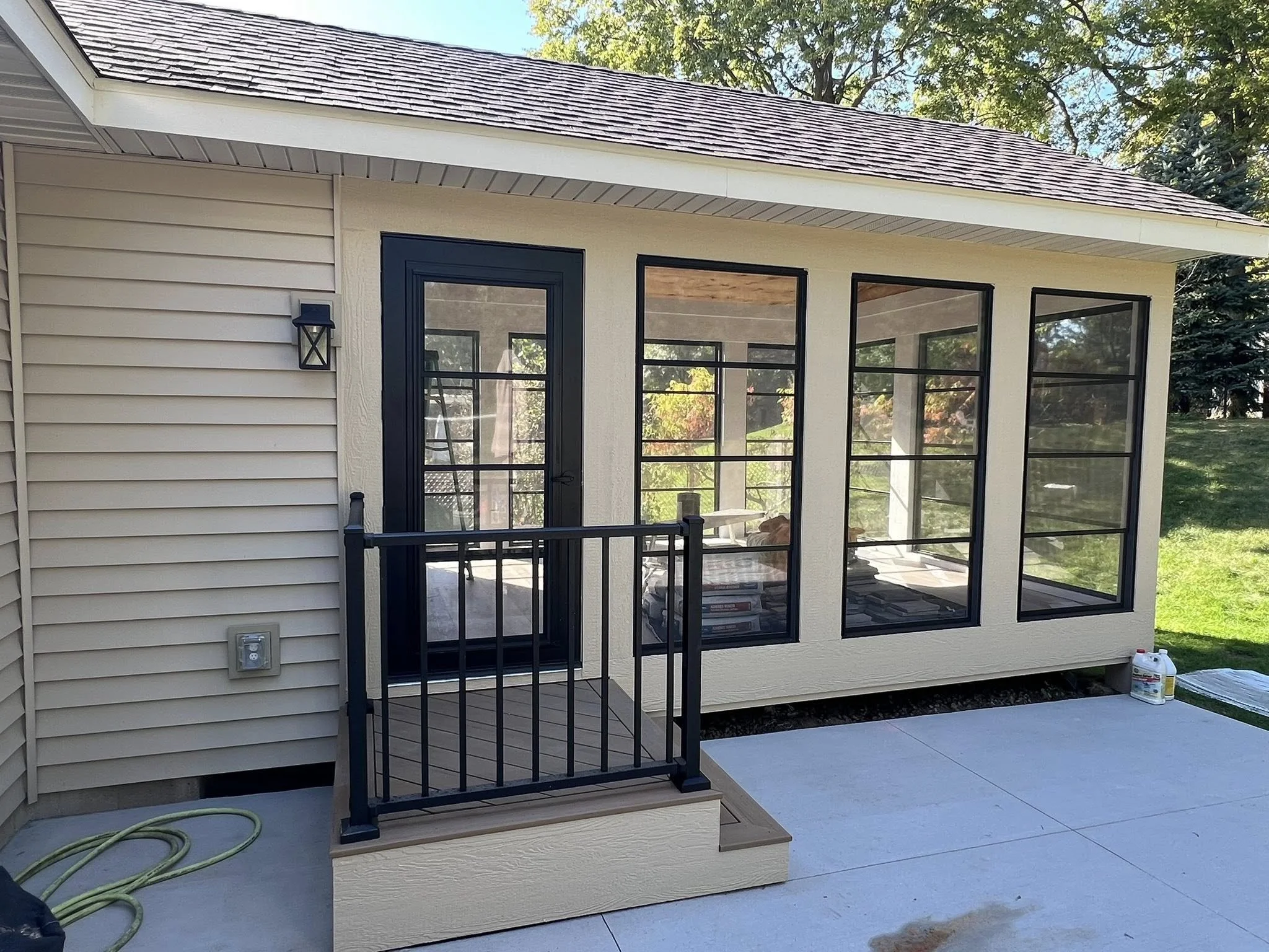 A newly built screened porch with large black-framed windows and a black door, attached to a beige house with vinyl siding, with a small set of stairs and a black railing, on a concrete slab with landscaping supplies nearby.