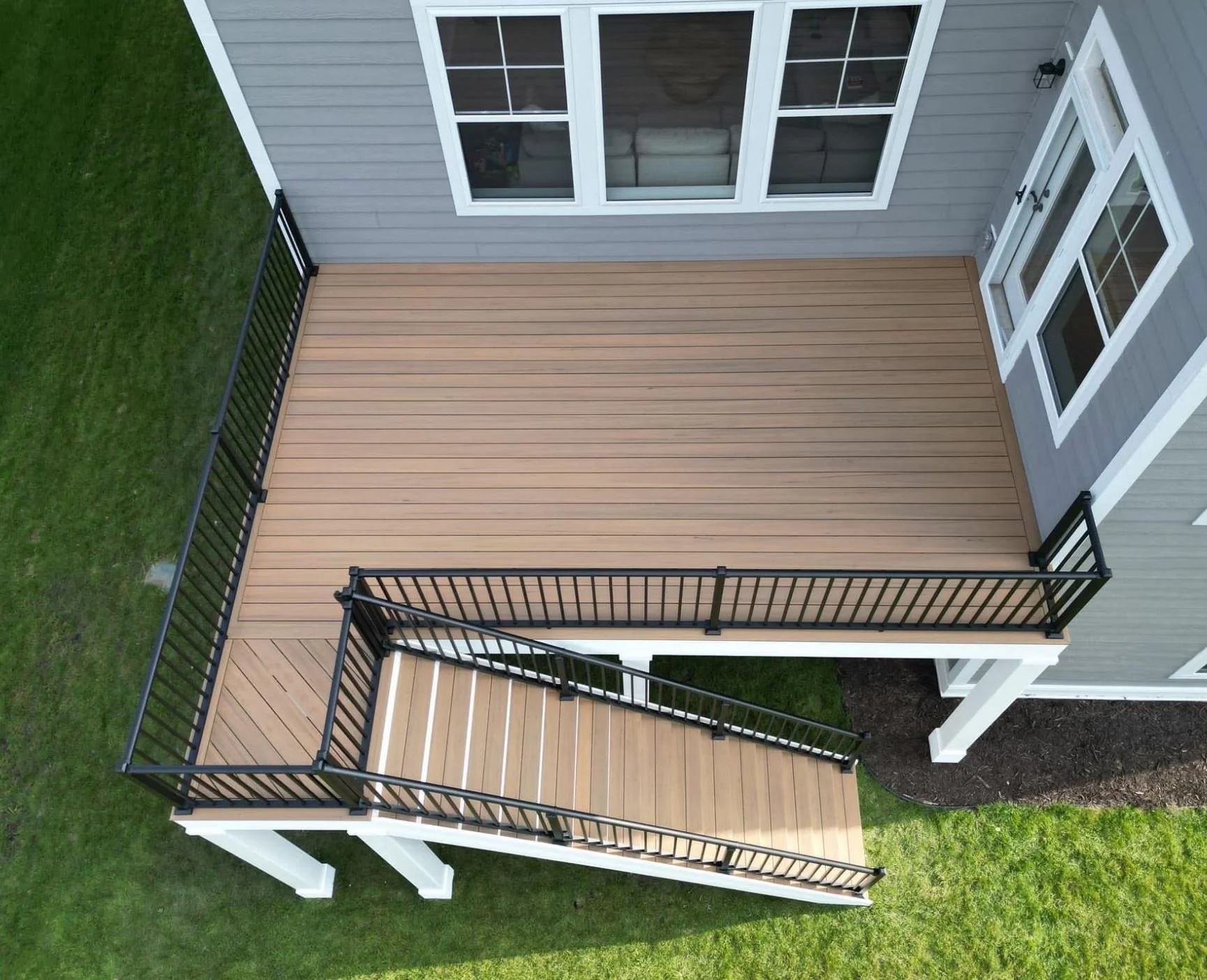 View of a newly built wooden deck attached to a gray house with white-trimmed windows and a glass door, enclosed by black metal railings, surrounded by a green lawn.