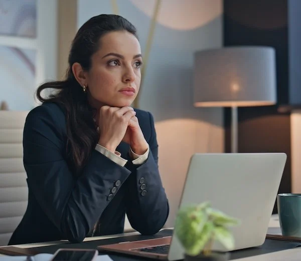 A professional woman in a dark blazer sits at a modern desk with her chin resting on her clasped hands, looking off-camera with a thoughtful or contemplative expression.
