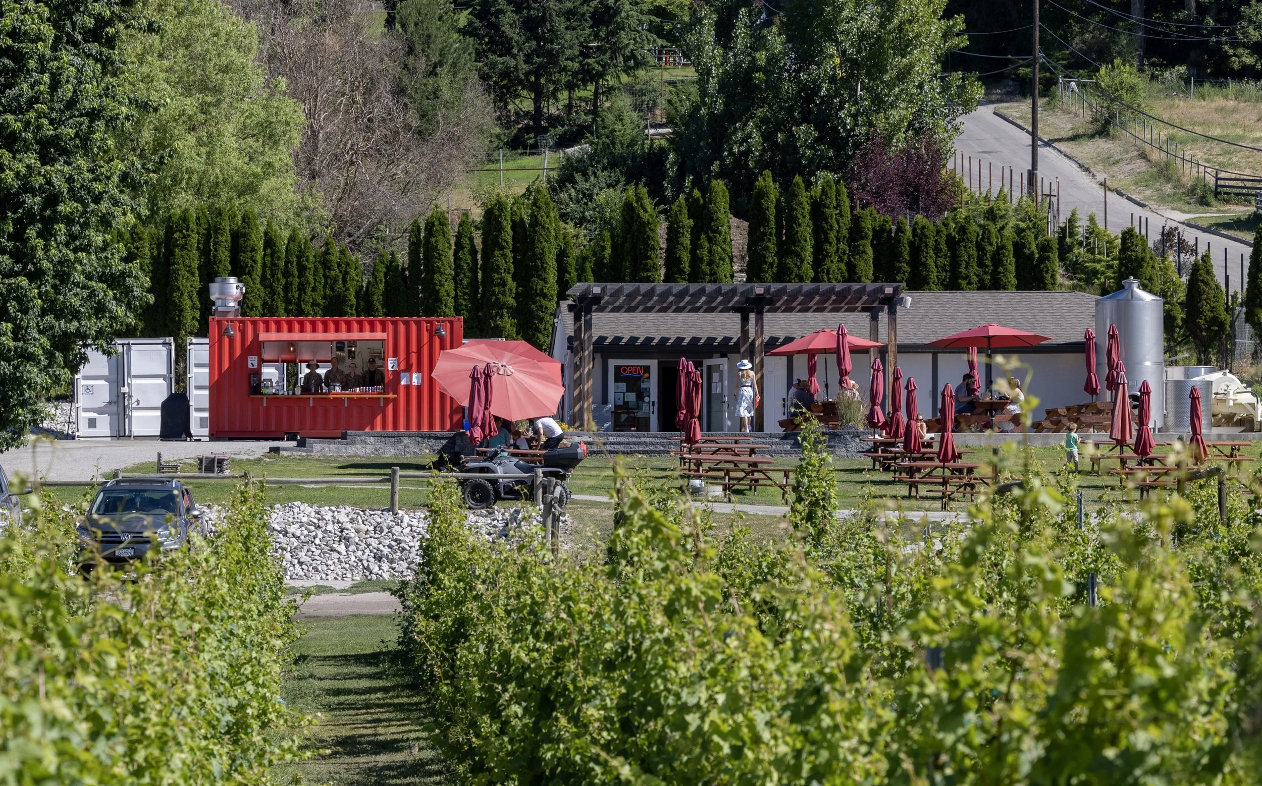 Outdoor seating area with red umbrellas and wooden benches in front of a building, surrounded by green trees and nearby road.