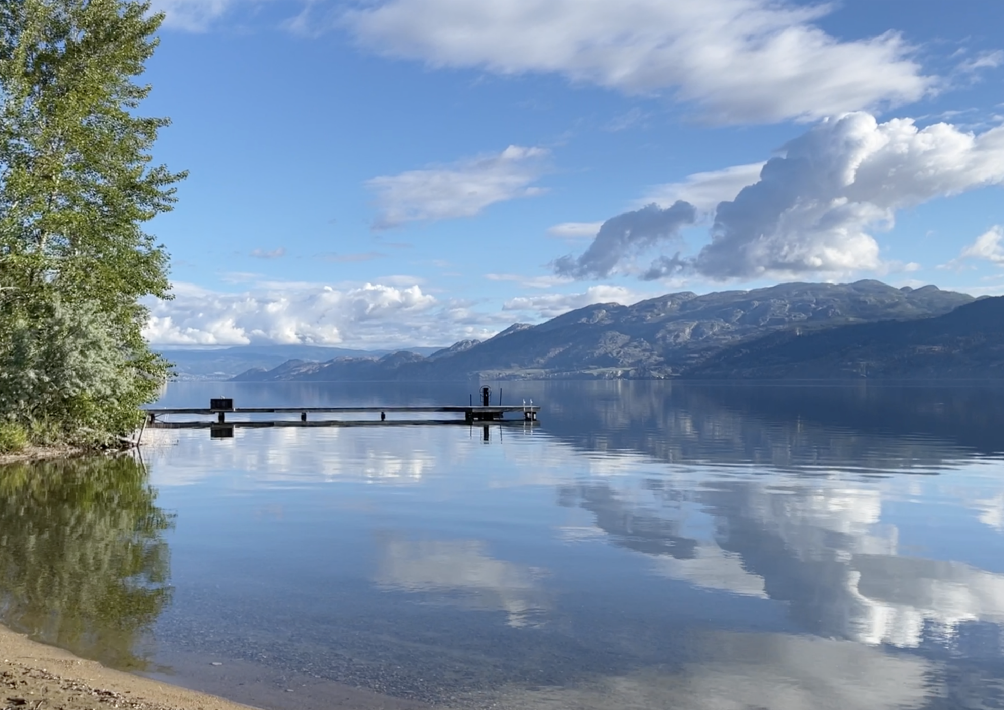 A scenic view of a lake with a dock, surrounded by trees and mountains under a partly cloudy sky.