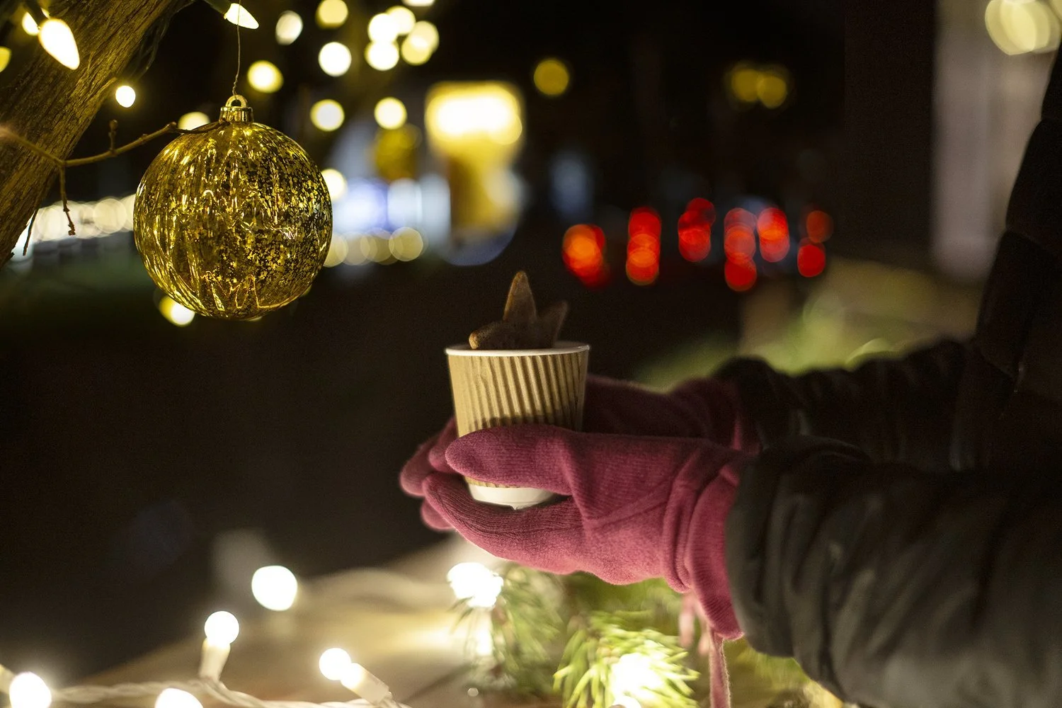 Person in a black jacket and pink gloves holding a cup with a hot drink, with a small animal's ear visible inside, near a decorated tree with a gold ornament and string lights at night.