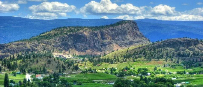 Scenic landscape with a mountain ridge, green hills, trees, and scattered houses under a partly cloudy sky.