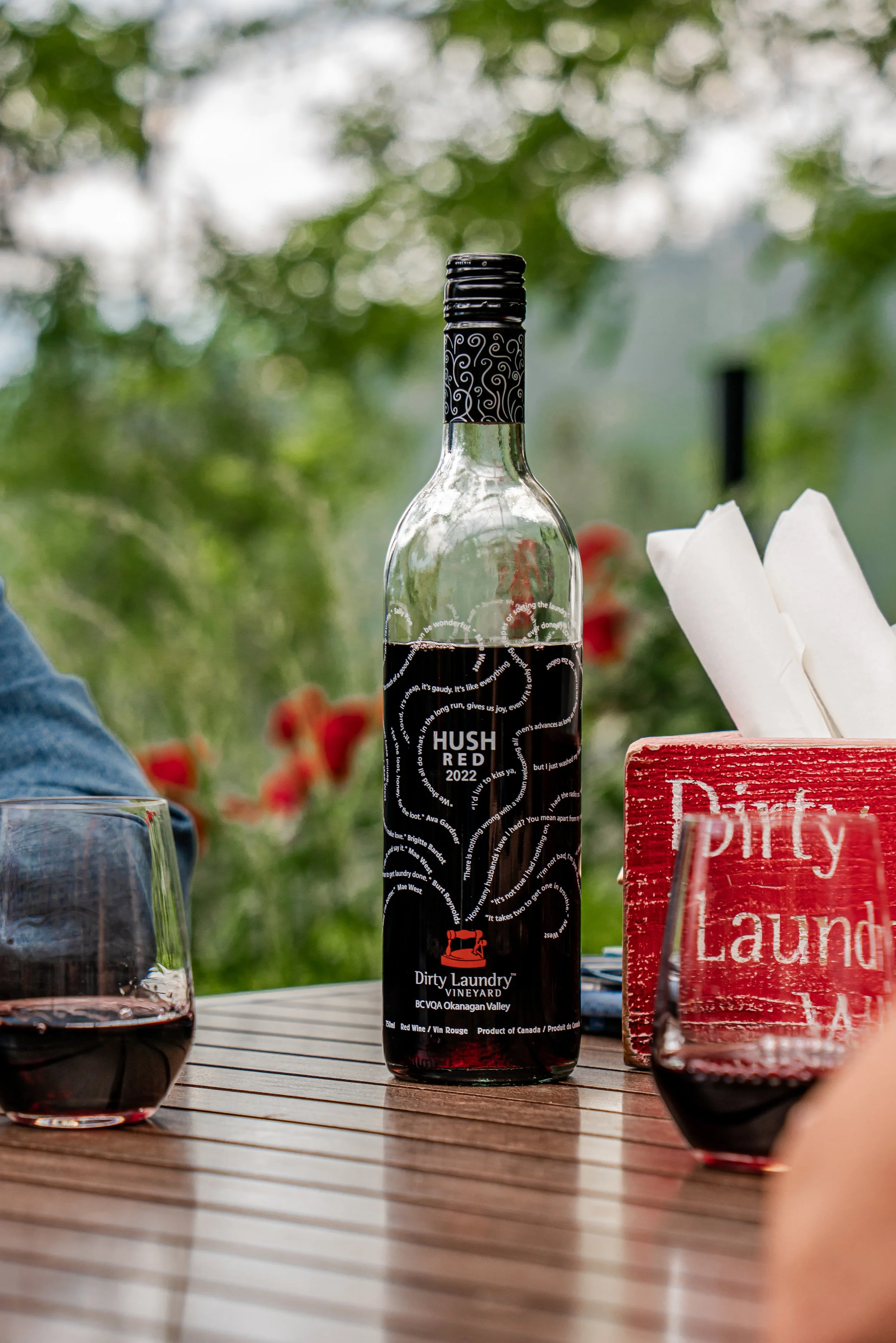 A bottle of Dirty Laundry Vineyard red wine set on a table, surrounded by glasses of red wine, with a red sign that reads 'Dirty Laundry' and a background of green foliage.