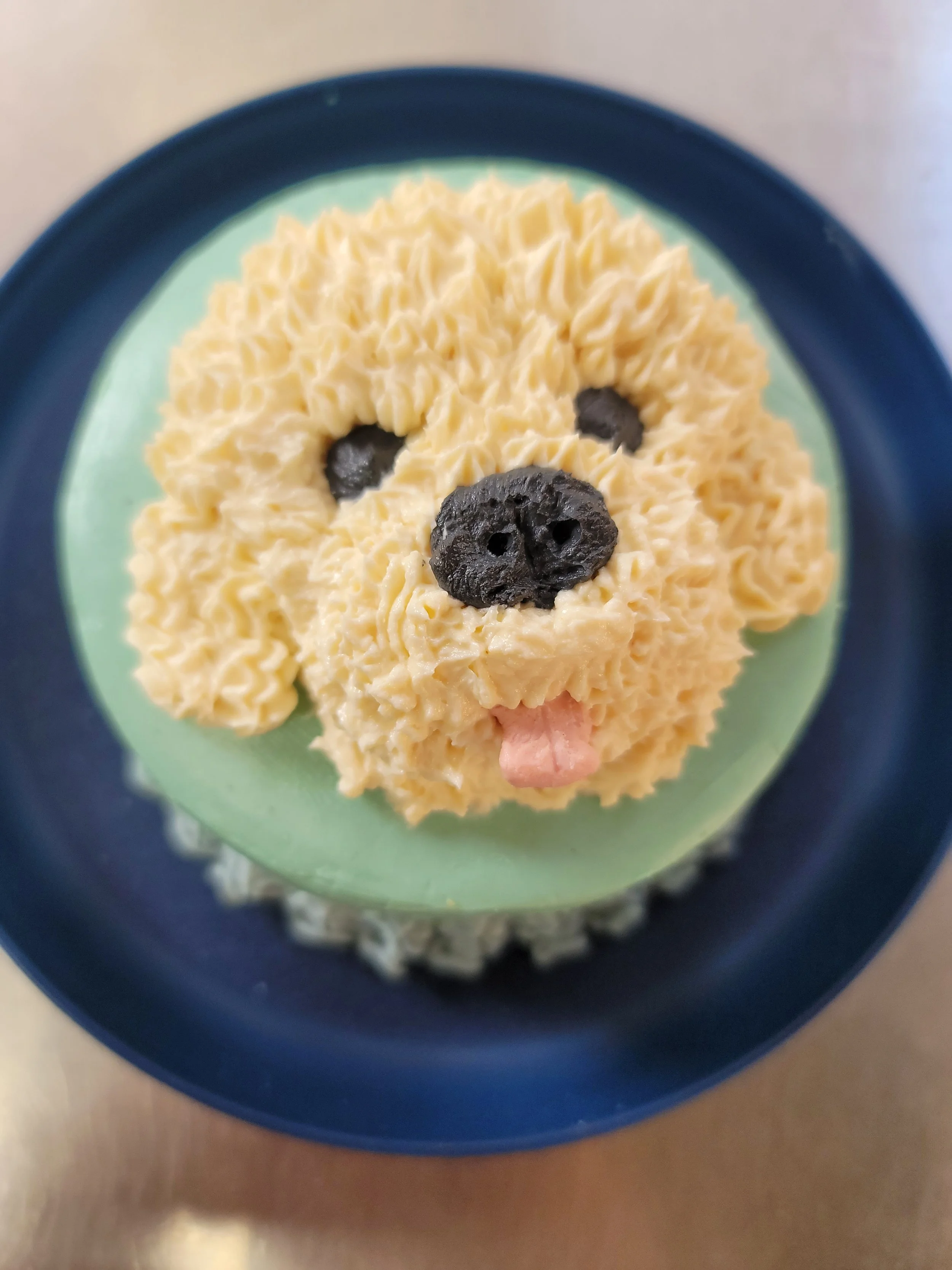 A cupcake with yellow frosting decorated to resemble a bear's face, with black candy for eyes and nose, on a green and blue plate.