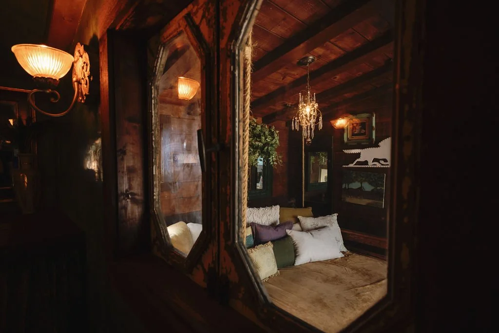 A cozy living room viewed through a vintage mirror with an ornate frame. The room features a wooden ceiling with exposed beams, a chandelier hanging from the ceiling, and framed artwork on the walls. Beneath the artwork are decorative pillows arranged on the floor, creating a comfortable seating area.