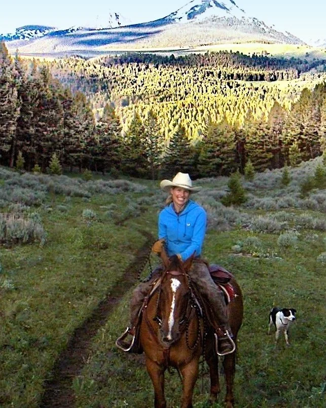 A woman wearing a cowboy hat and a blue jacket is riding a brown horse on a trail in a lush green landscape. A black and white dog walks beside her. In the background, there are dense evergreen trees, rolling hills, and snow-capped mountains under a partly cloudy sky.