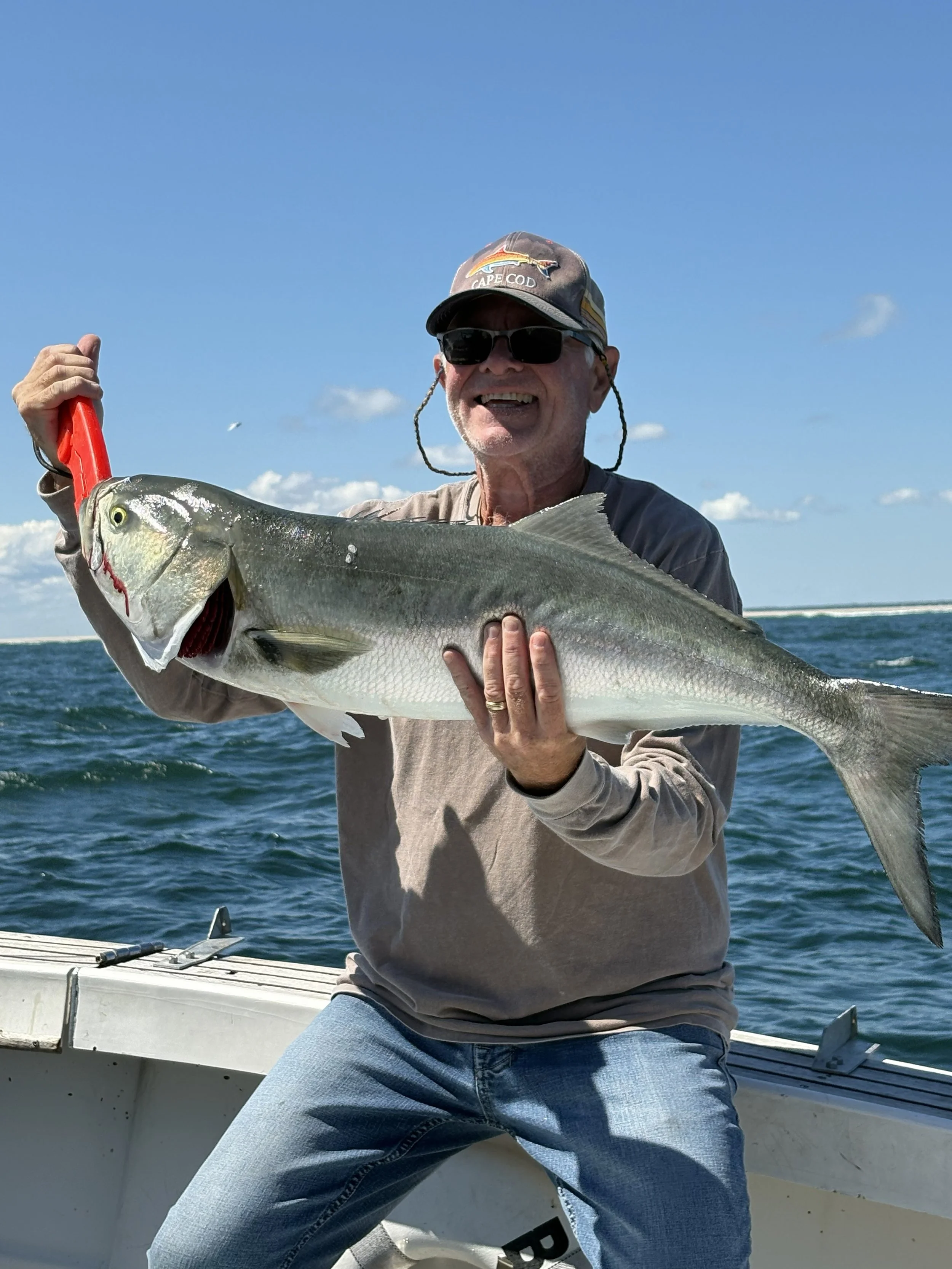 An older man wearing sunglasses, a cap, a gray shirt, and jeans, smiling while holding a large fish with a fishing tool, on a boat in the water under a blue sky with scattered clouds.