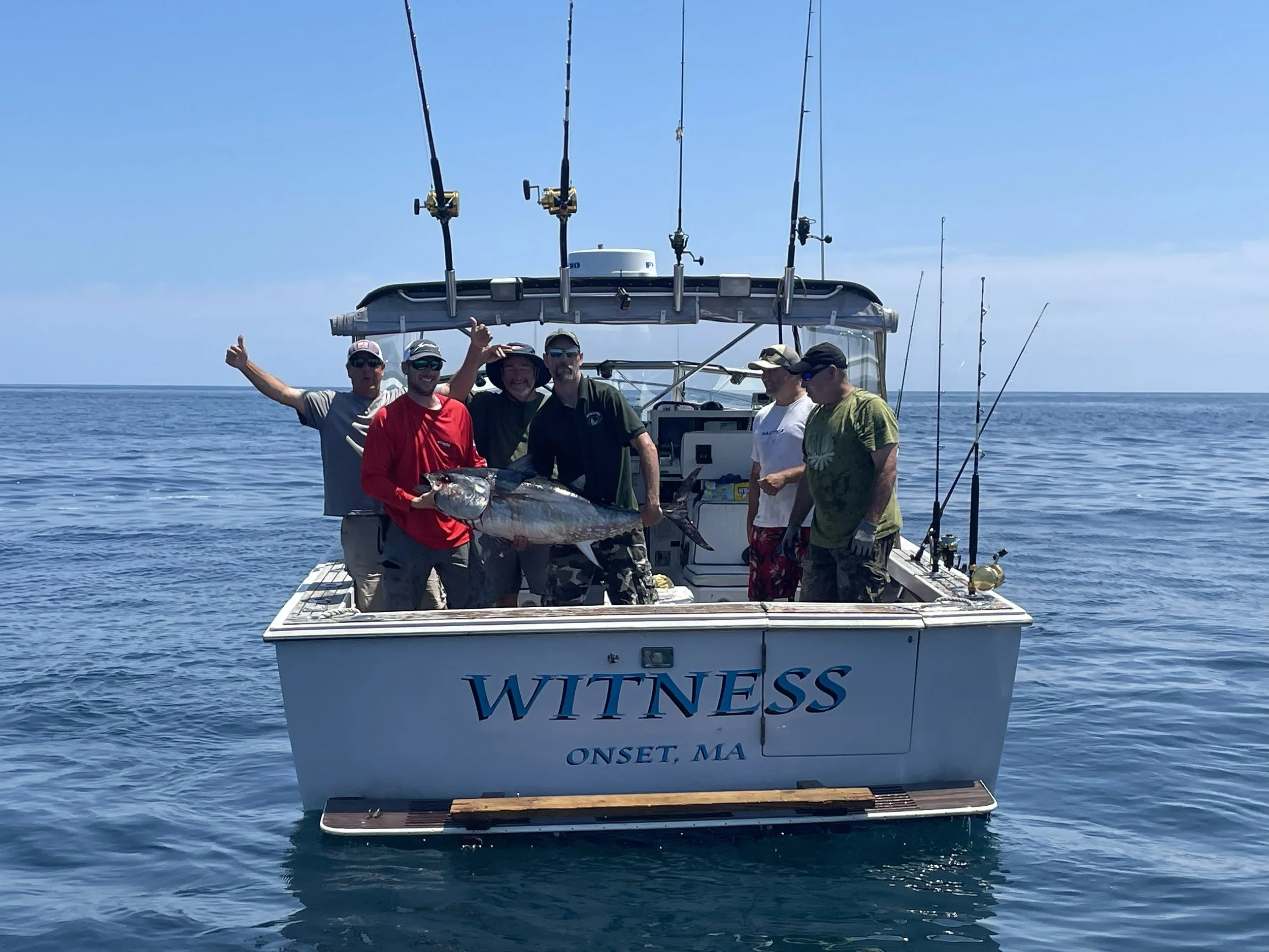Group of five men on a boat holding a large fish, with fishing rods mounted above them, in a body of water under a clear blue sky, with the boat displaying the words "Witness" and "Onset, MA."