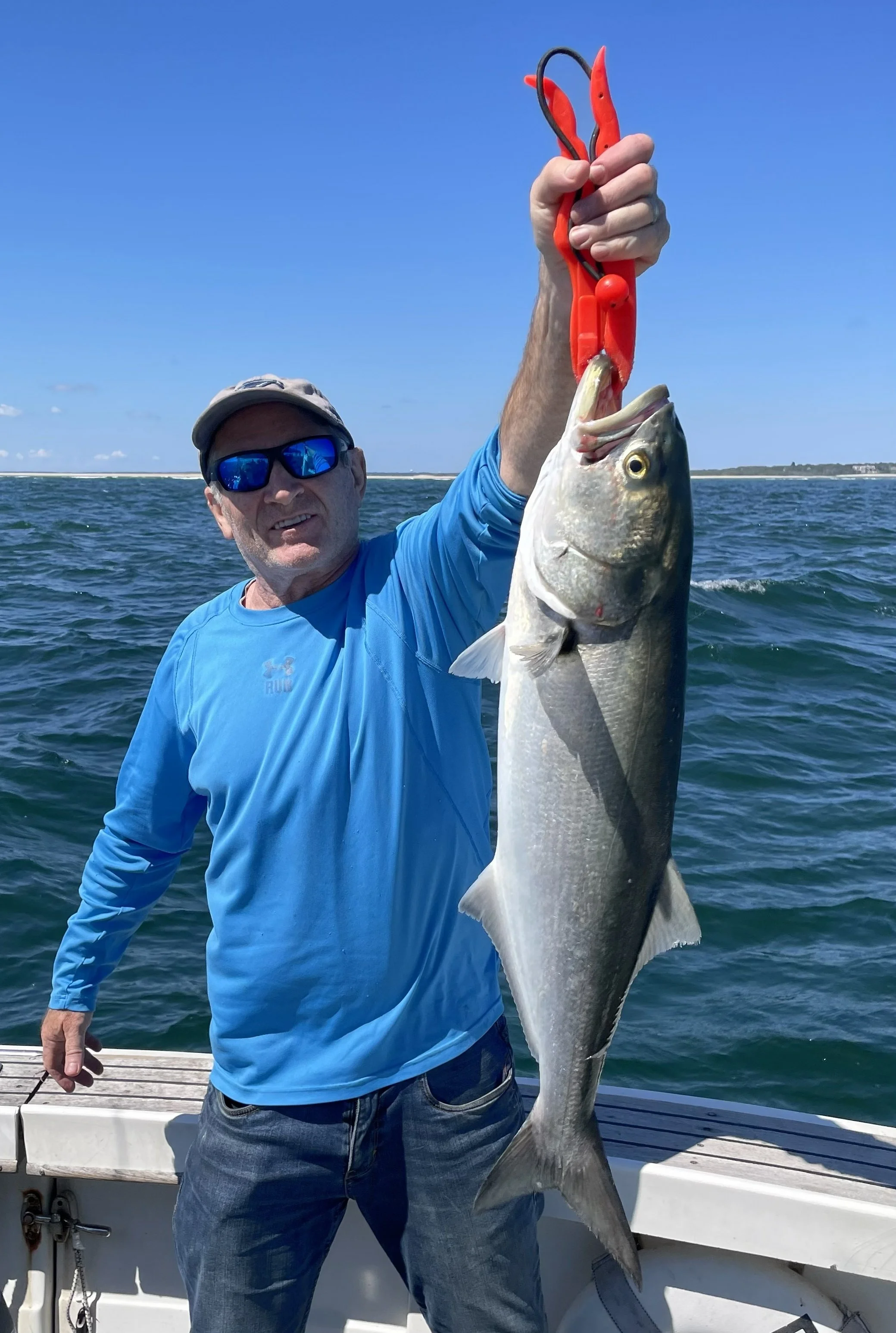 A man wearing a blue long-sleeve shirt, sunglasses, and a cap on a boat, holding a large fish with red fishing pliers against a backdrop of ocean and blue sky.