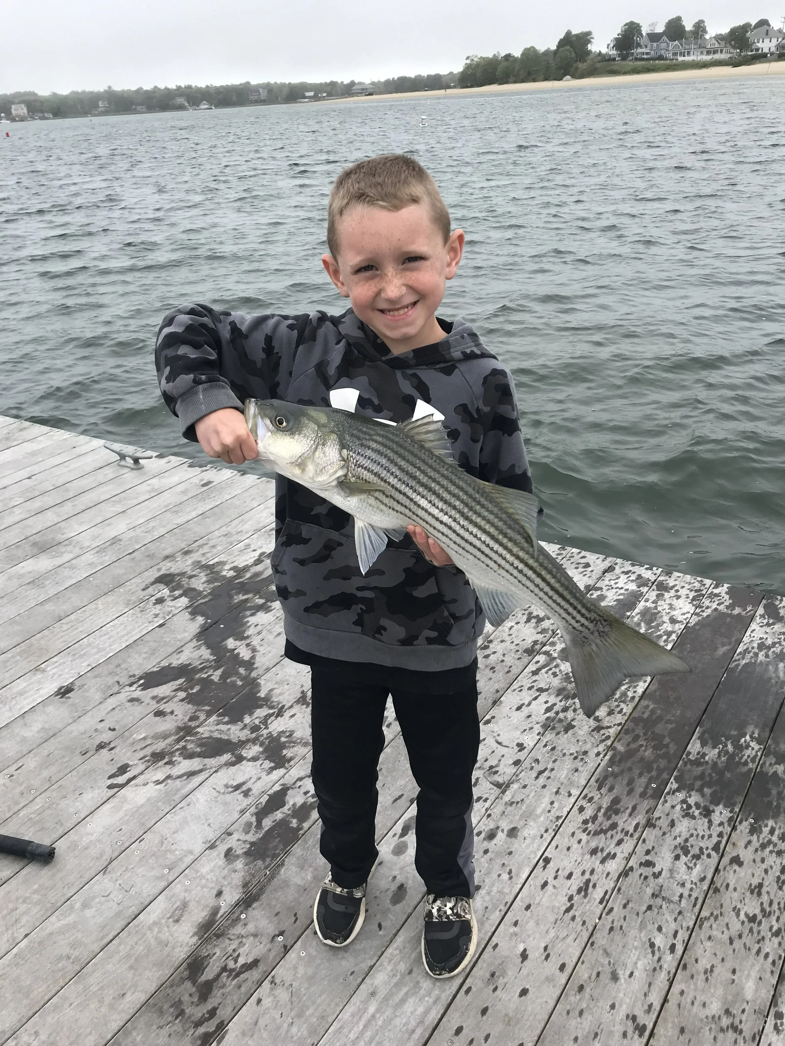 A young boy standing on a wooden dock at a lake, smiling and holding a large fish he caught.