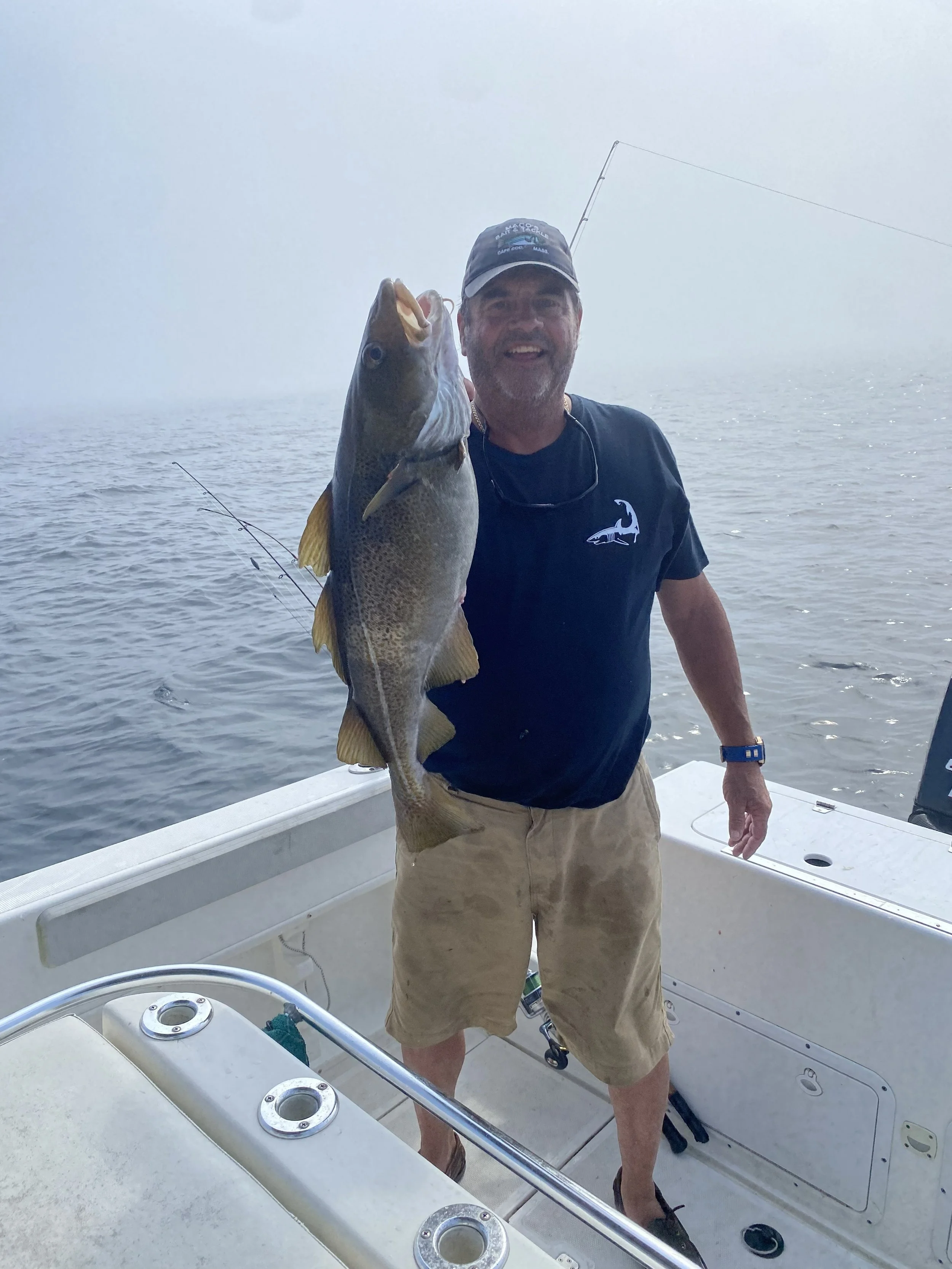 A man on a boat holding a large fish he caught, smiling at the camera, with water and foggy sky in the background.