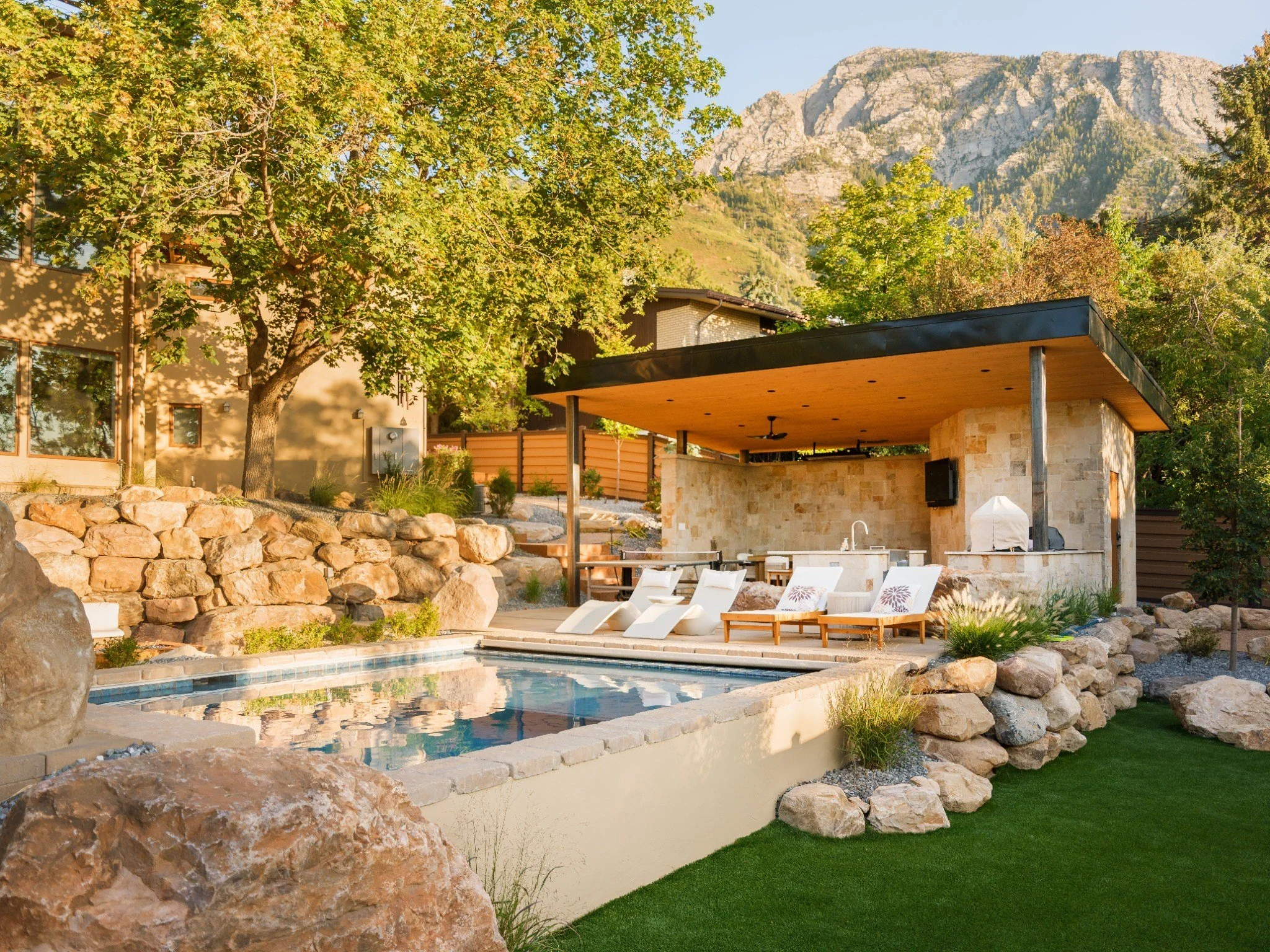 Utah backyard landscape design with swimming pool, natural boulder retaining walls, covered outdoor kitchen, lounge seating, and mountain views.