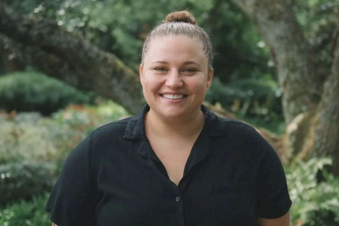 A woman with light skin and light hair in a bun, smiling, outdoors in front of green trees and bushes, wearing a black shirt.