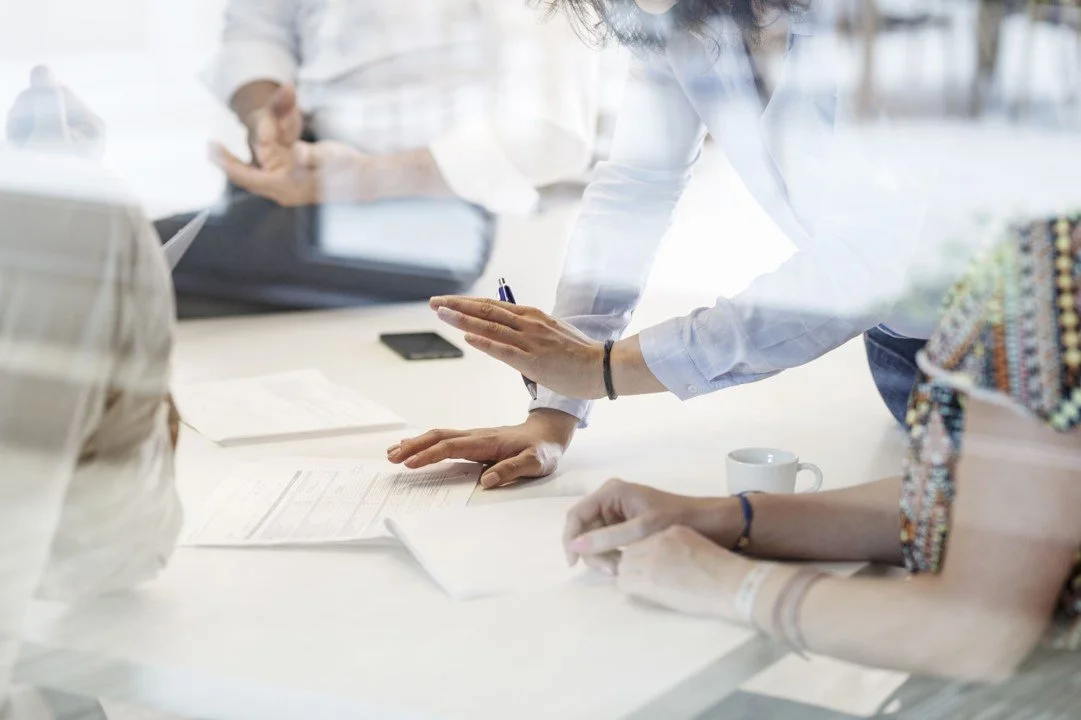 People in a meeting room discussing documents with a laptop, tablet, and coffee cup on the table.