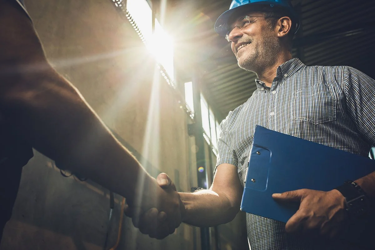 Two men shaking hands in an industrial setting, with one holding a blue clipboard, sunlight shining through a window.