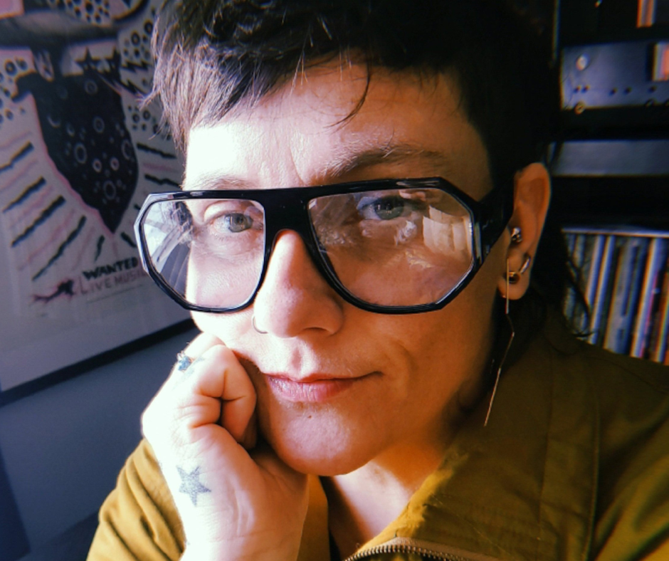 Close-up of a person wearing large black glasses, with short dark hair, resting their chin on their hand, with a star tattoo on their hand, in a room with bookshelves and a colorful poster in the background.
