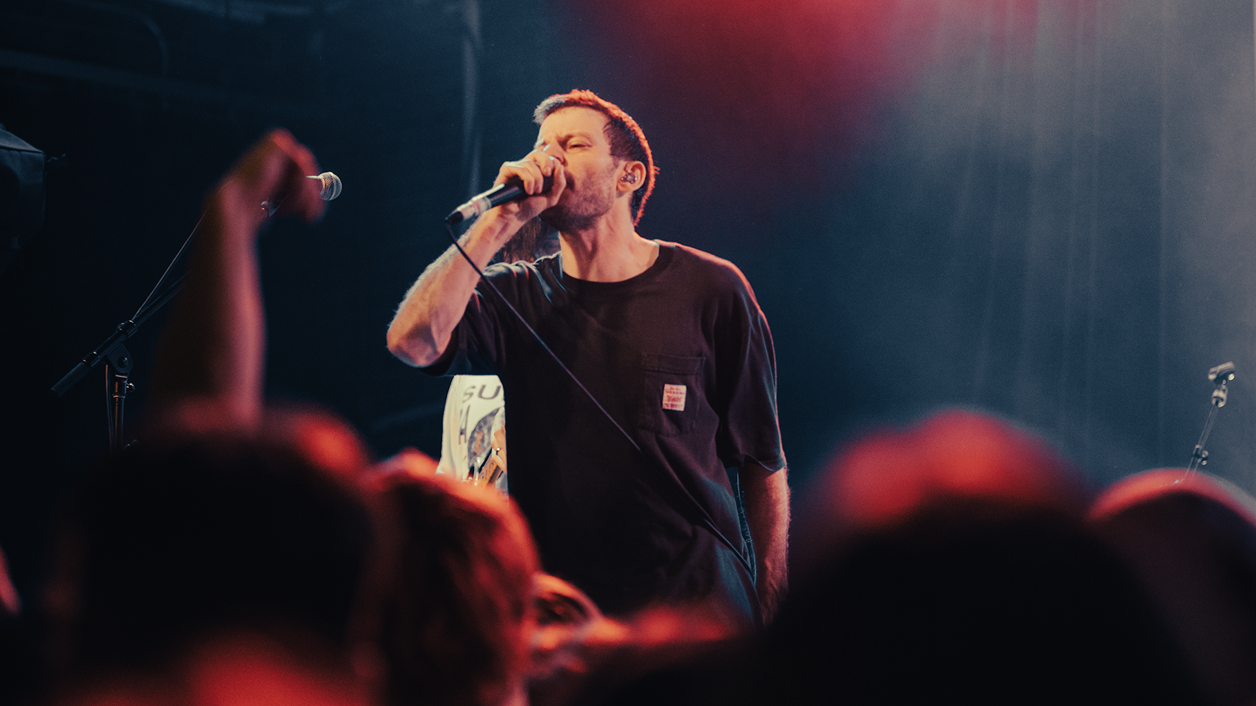 A male singer with short hair and a beard singing into a microphone on a stage, wearing a black t-shirt during a concert. The stage is dimly lit with colored lights, and there are microphones and people in the audience visible in the foreground.