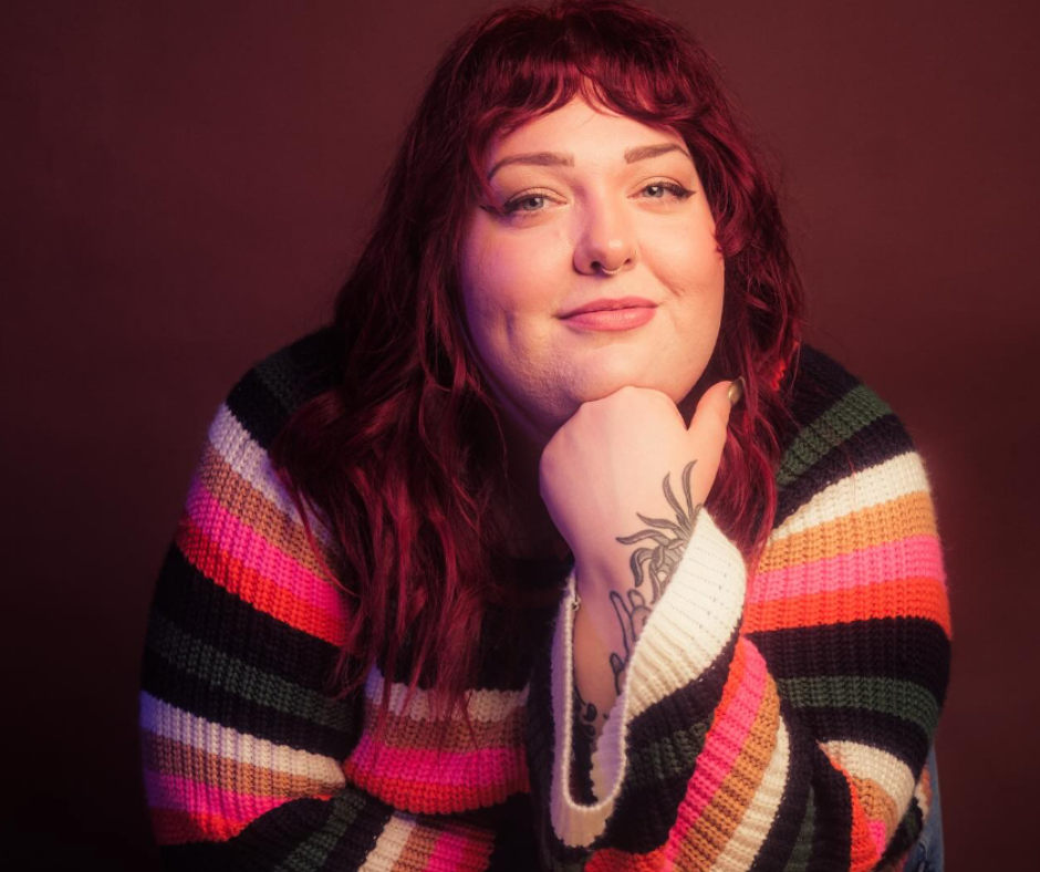 A woman with shoulder-length wavy red hair and light skin, wearing a multicolored striped sweater, resting her chin on her hand with a tattoo visible on her wrist, against a dark pink background.