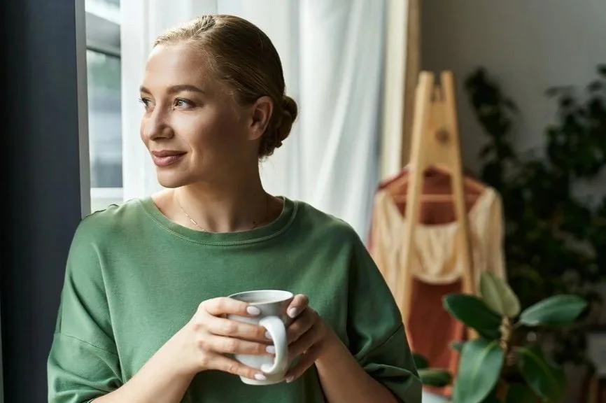 Woman holding a warm mug beside a window surrounded by natural light and indoor plants, representing wellbeing, rest and recovery.