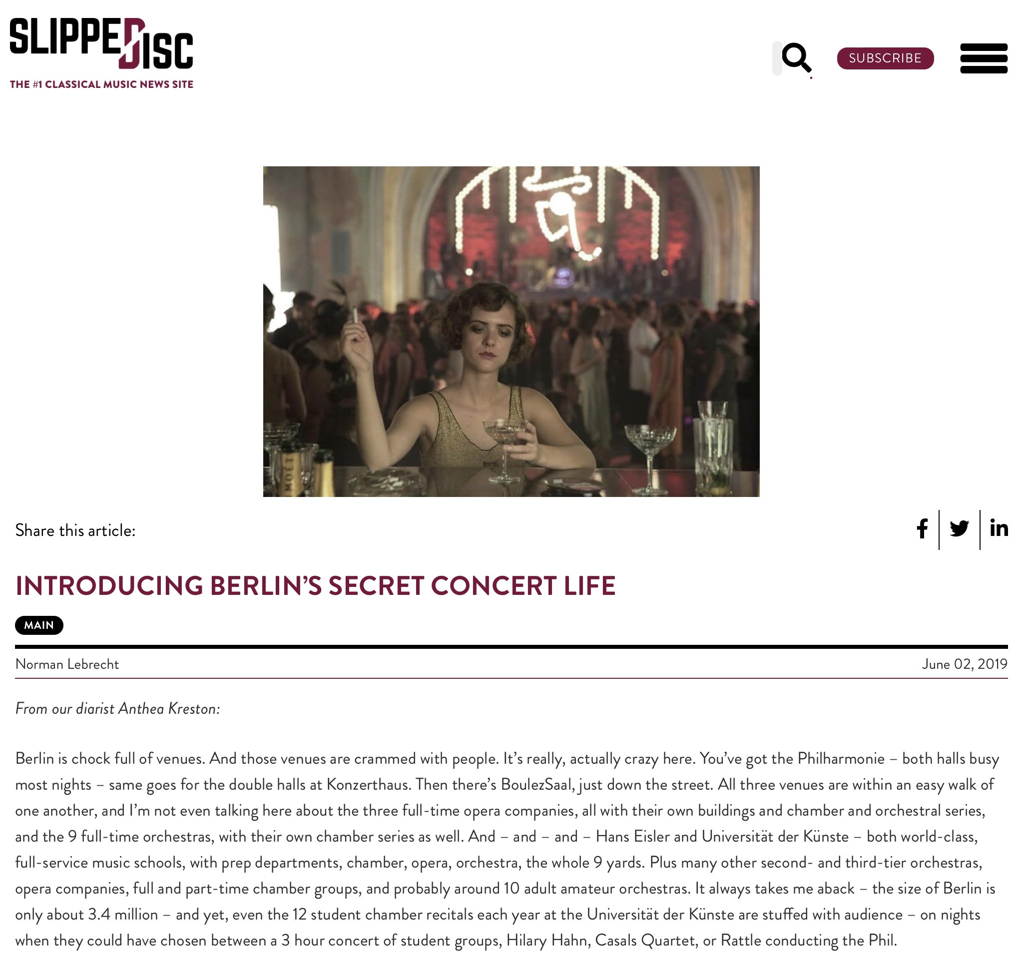 A woman with short wavy hair in a gold dress, sitting at a bar in a crowded theater with a large, ornate arch and a neon sign in the background. She is holding a cocktail glass and has a calm, composed expression.