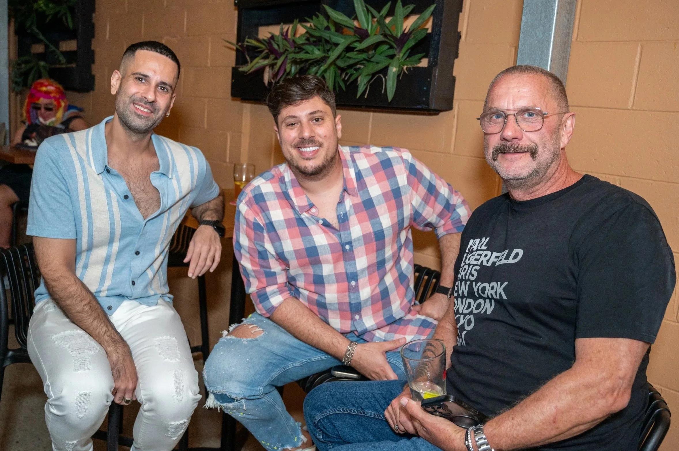 Three men sitting together at a social gathering, smiling at the camera. The man on the left is wearing a light blue button-up shirt and white ripped jeans. The man in the middle is wearing a pink and blue plaid shirt and ripped jeans. The man on the right is wearing glasses and a black t-shirt with white text. There is a woman in the background with colorful hair, sitting at a table. The setting appears to be indoors with warm-colored walls and decorative plants.