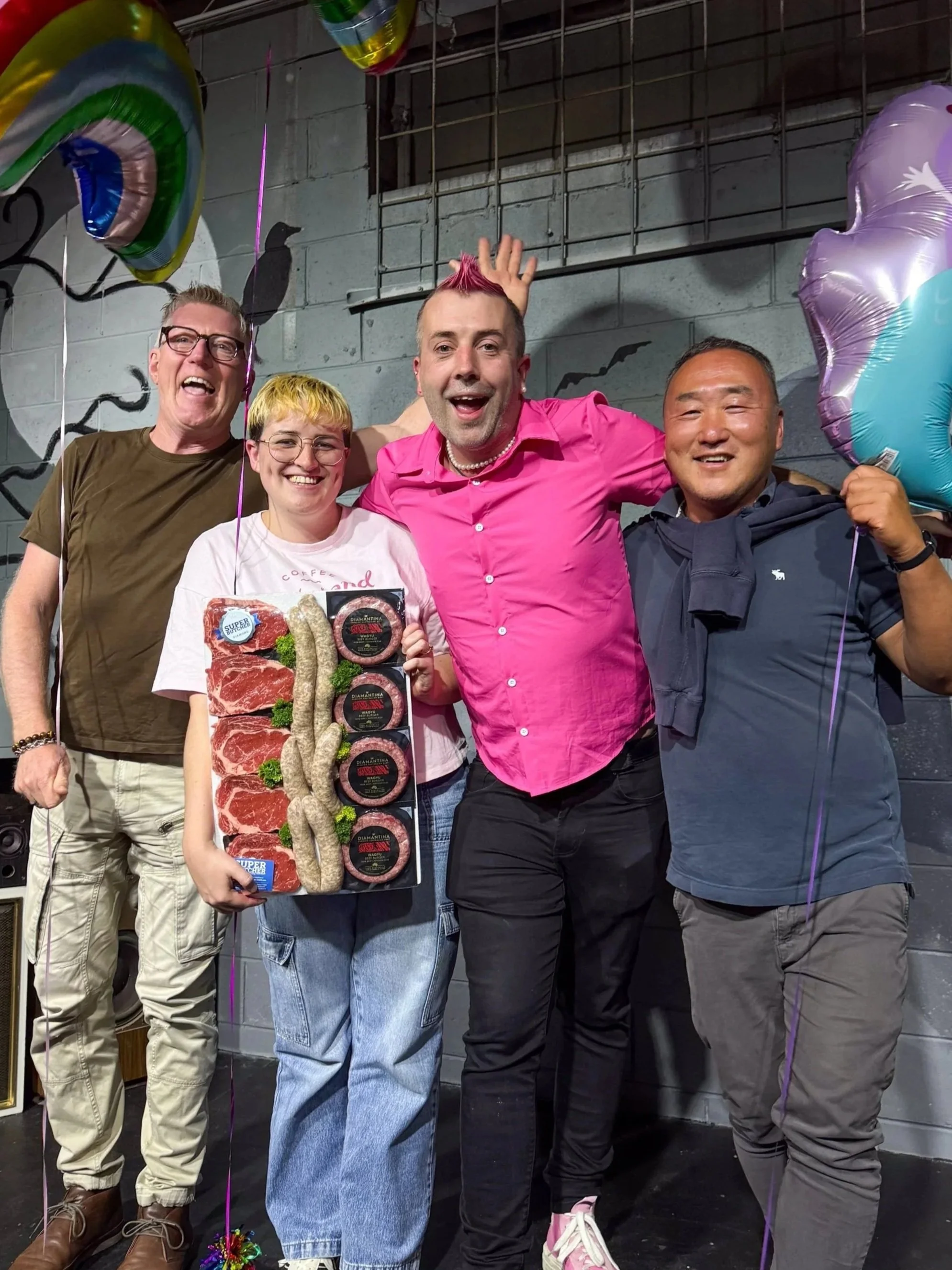 A group of four people celebrating together at a party, with balloons and festive decorations. The person second from the right has pink hair and is wearing a pink shirt, and is holding a tray of food including meats and sausages. They are smiling and appear to be happy.