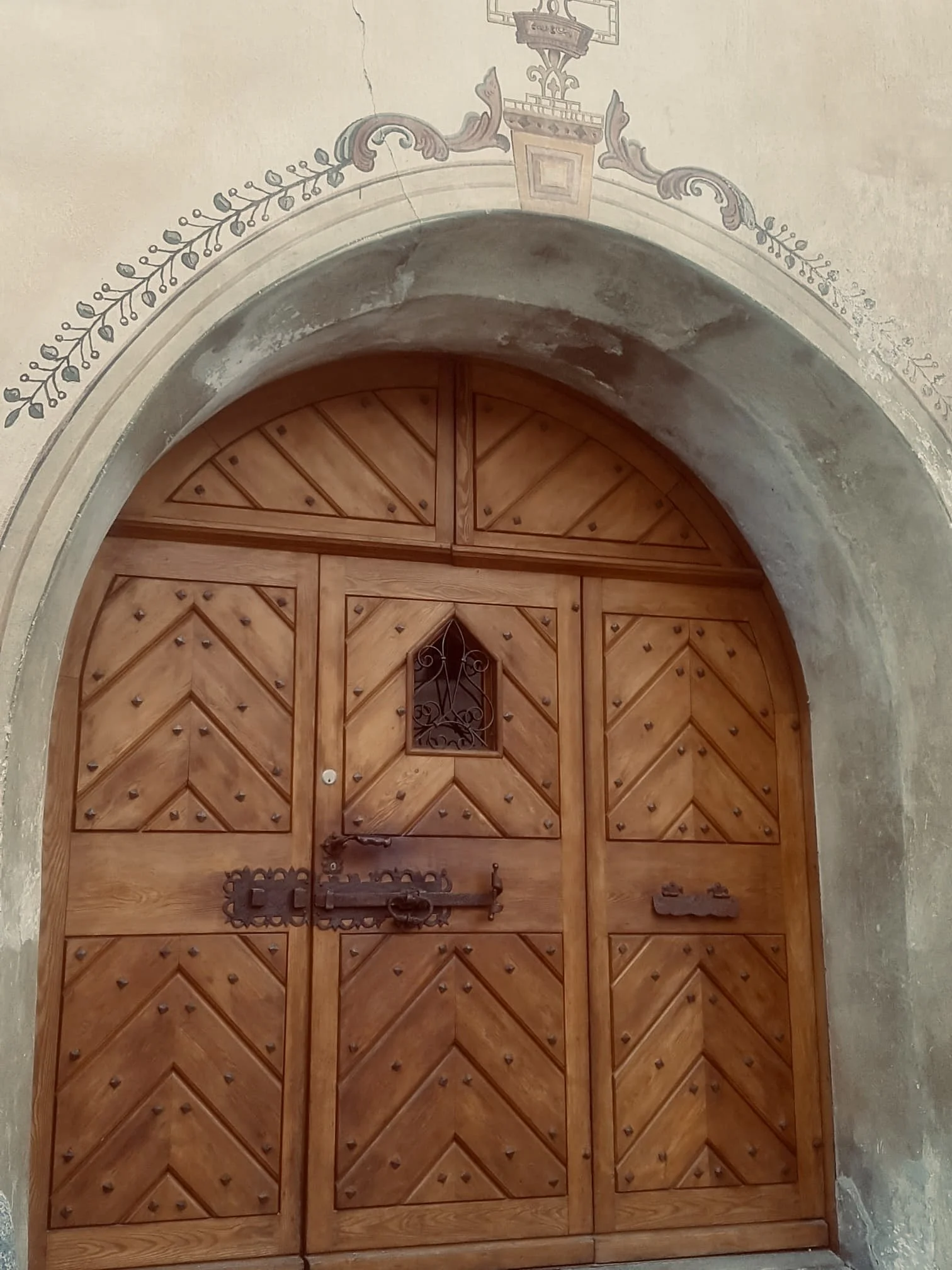 Ein geschnitztes Holztür mit kleinen Metallakzenten und einem kleinen, schmiedeeisernen Gitterfenster im oberen Bereich, umgeben von einer alten, verputzten Wand mit Wandmalerei und einem Rundbogen