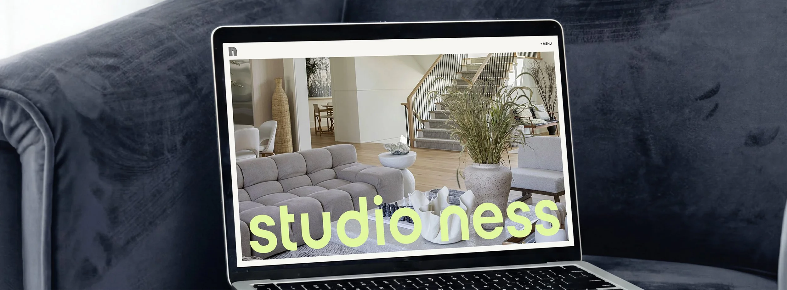 Laptop displaying a photo of a modern living room with a gray couch, leafy plant, white decorative items, and a staircase in the background.