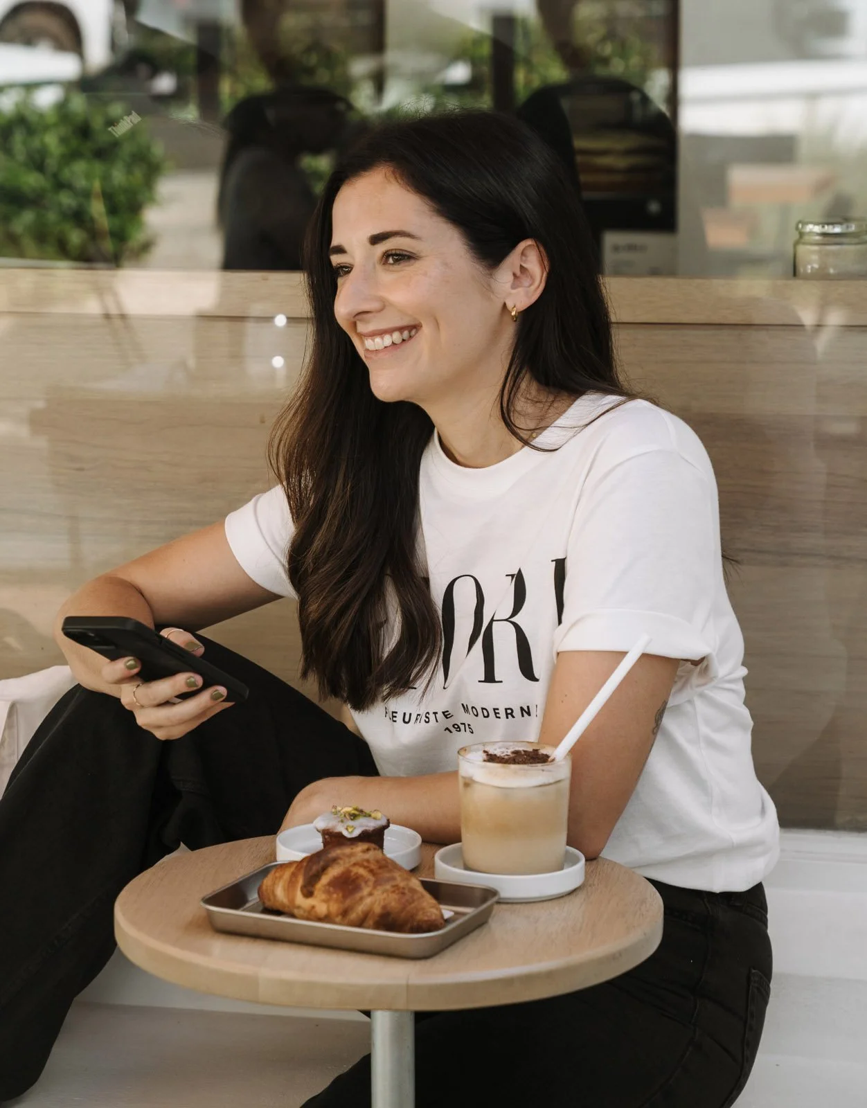 A woman with long dark hair, wearing a white t-shirt with black text, sitting at a table in a cafe, smiling while looking at her phone. On the table, there is a tray with a croissant, a small dessert, and a glass of coffee.