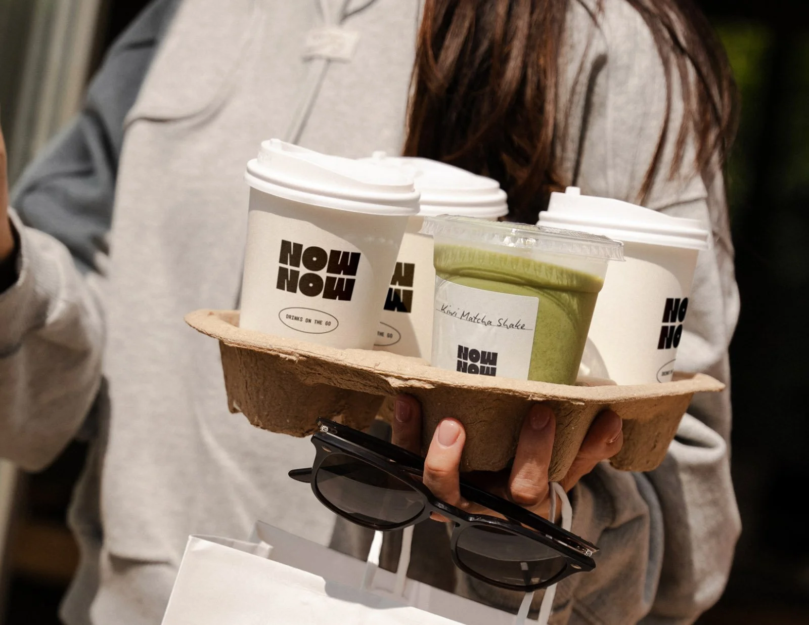A person holding a paper tray with three coffee cups and a kiwi matcha shake, with a pair of sunglasses hanging from their hand.