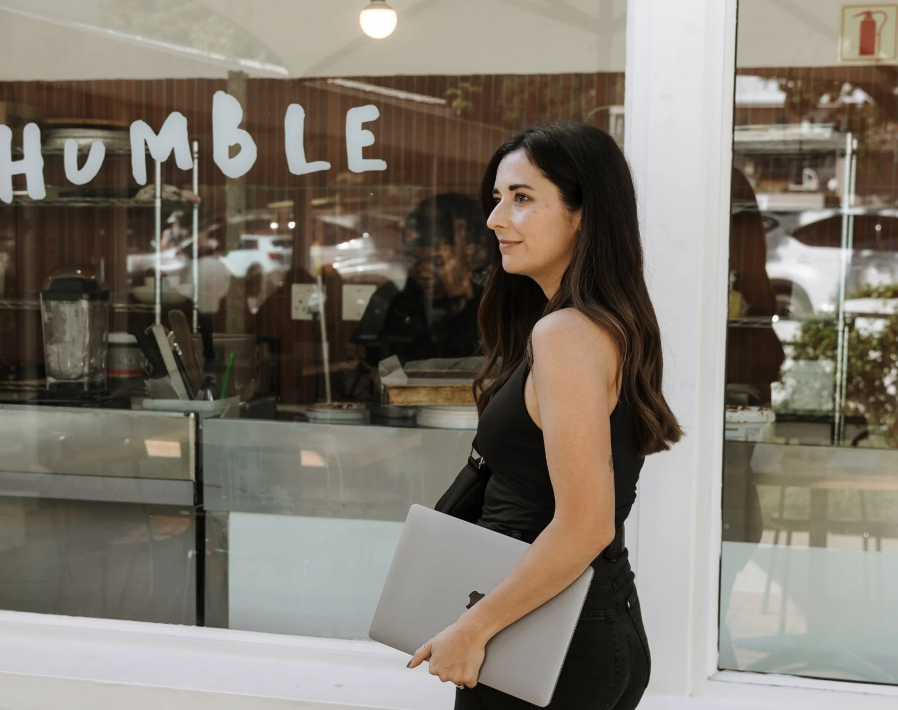 A woman with long dark hair and light skin holds a laptop and a folder in front of a glass window, with a reflection of the street outside.