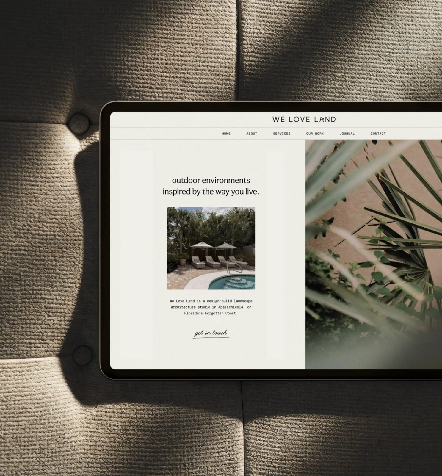 Part of a tablet showing a website for a landscape architecture studio called We Love Land, featuring outdoor environments and a poolside scene, placed on a textured woven beige surface with shadows.