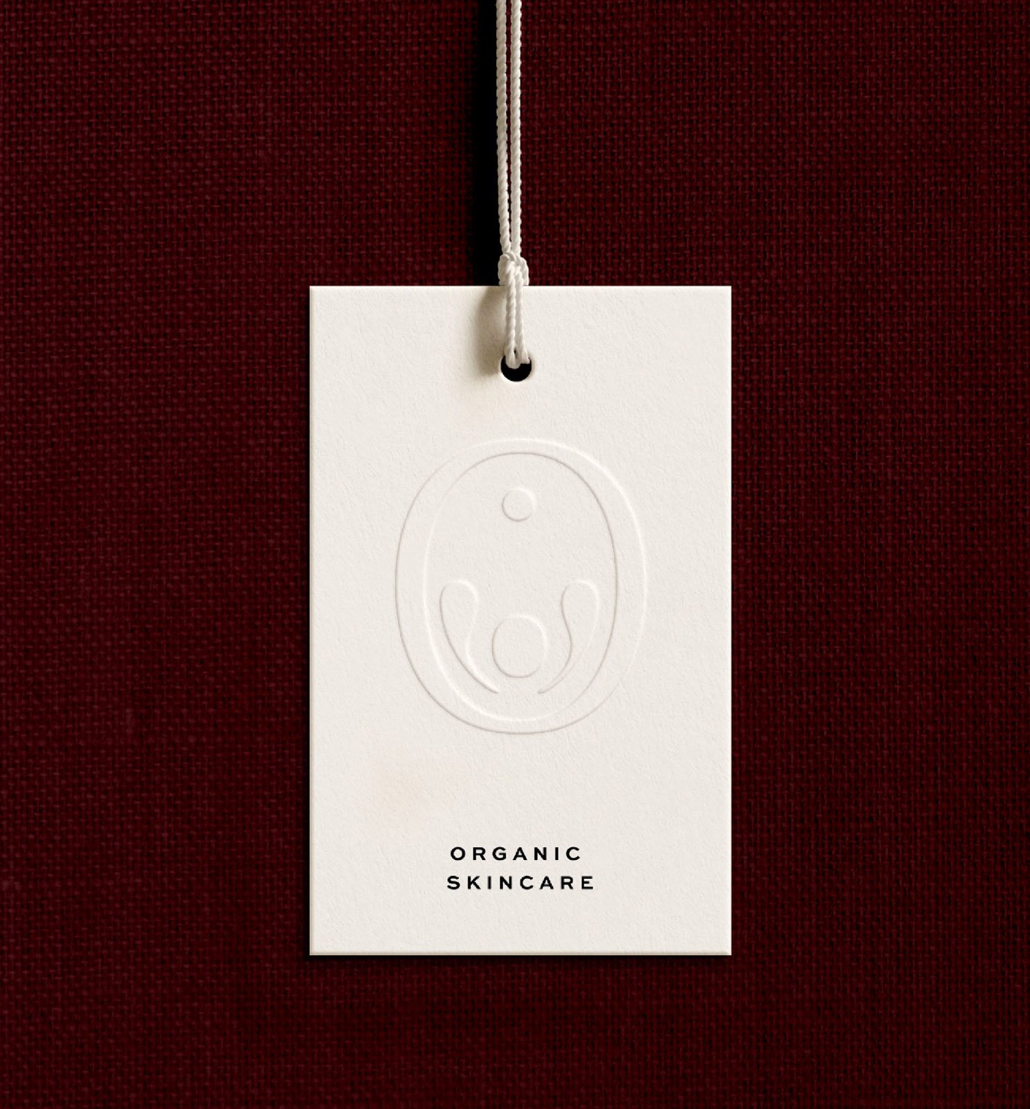 A white skincare product tag with a minimalist embossed face logo hanging from a white string, set against a dark red textured background, with the text 'Organic Skincare' at the bottom.