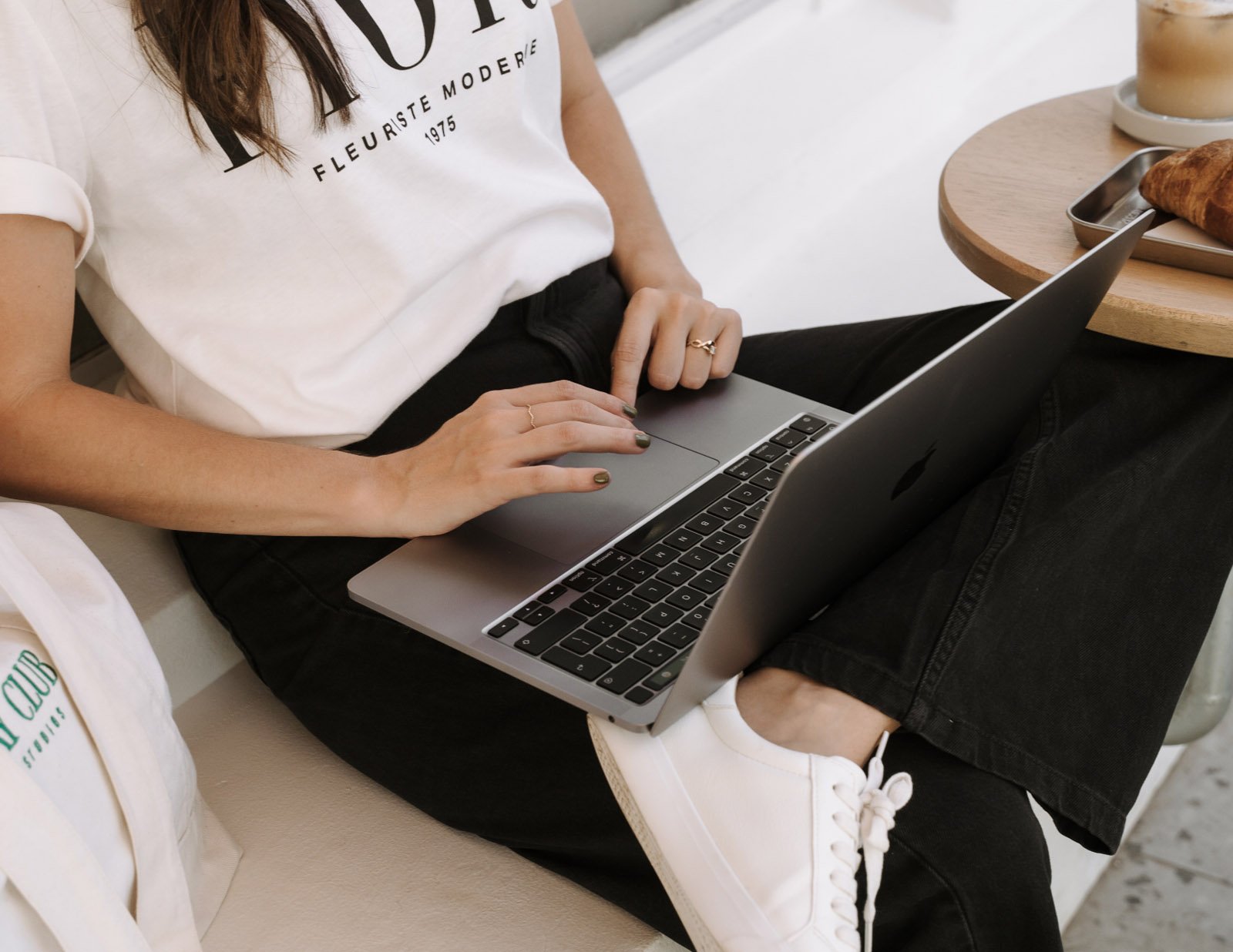 Person using a silver MacBook laptop while sitting on a beige couch, wearing black pants, white sneakers, a white T-shirt, and jewelry, with a round wooden table nearby holding a tray with a baked croissant and a beverage.