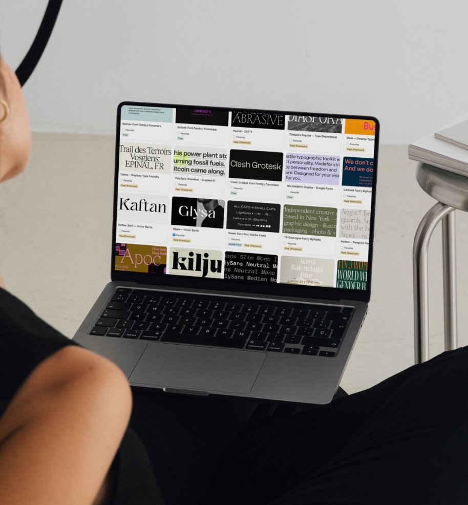 Person using a MacBook laptop with a display showing various font and typeface samples and names, in a modern workspace with a white wall and a chair.