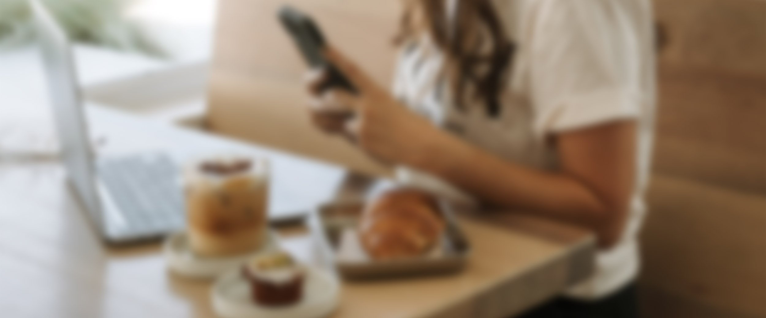 A person using a smartphone at a table with a tray of cupcakes, a glass of iced coffee, and a laptop.