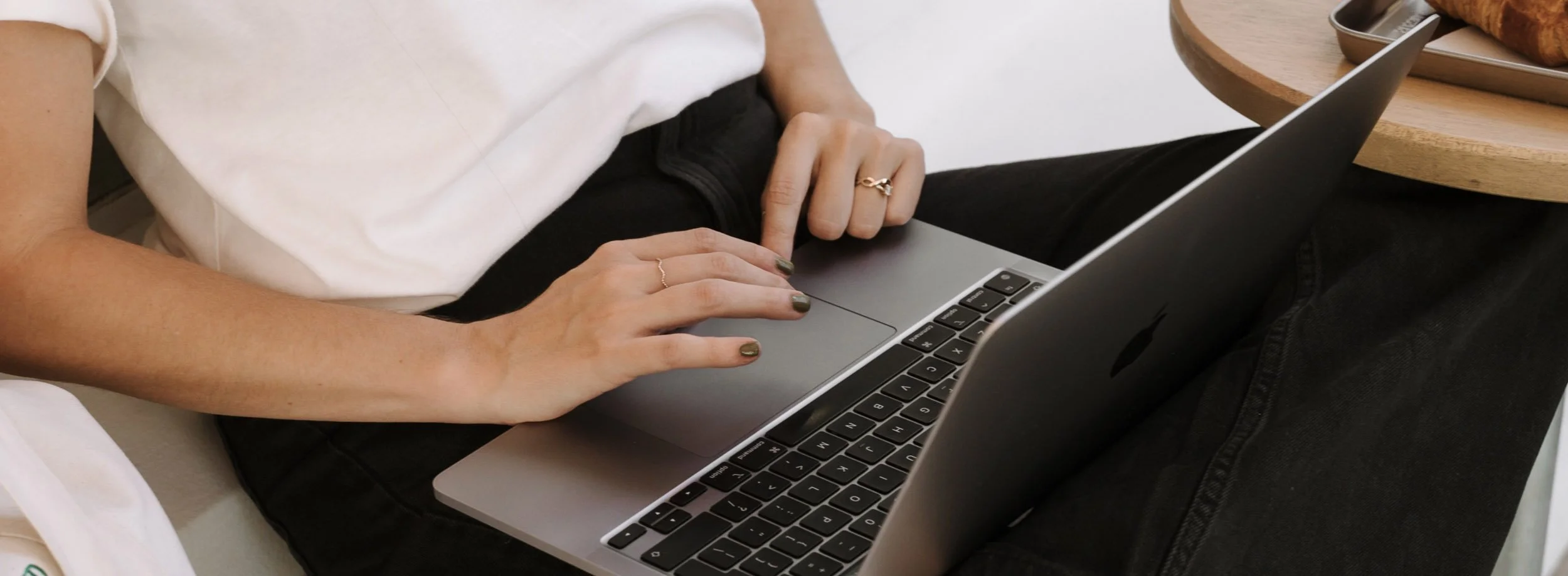 Person typing on a silver laptop with a black keyboard, sitting on a beige couch near a wooden table.