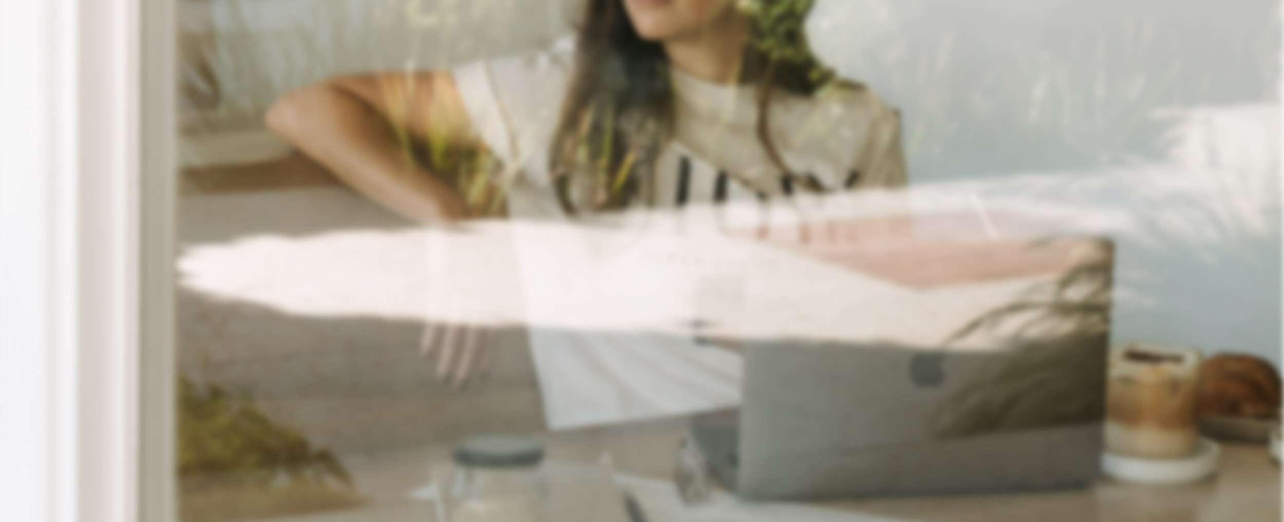 A woman with wavy hair sitting at a table with a laptop, papers, and a coffee cup, seen through a glass window with reflections.