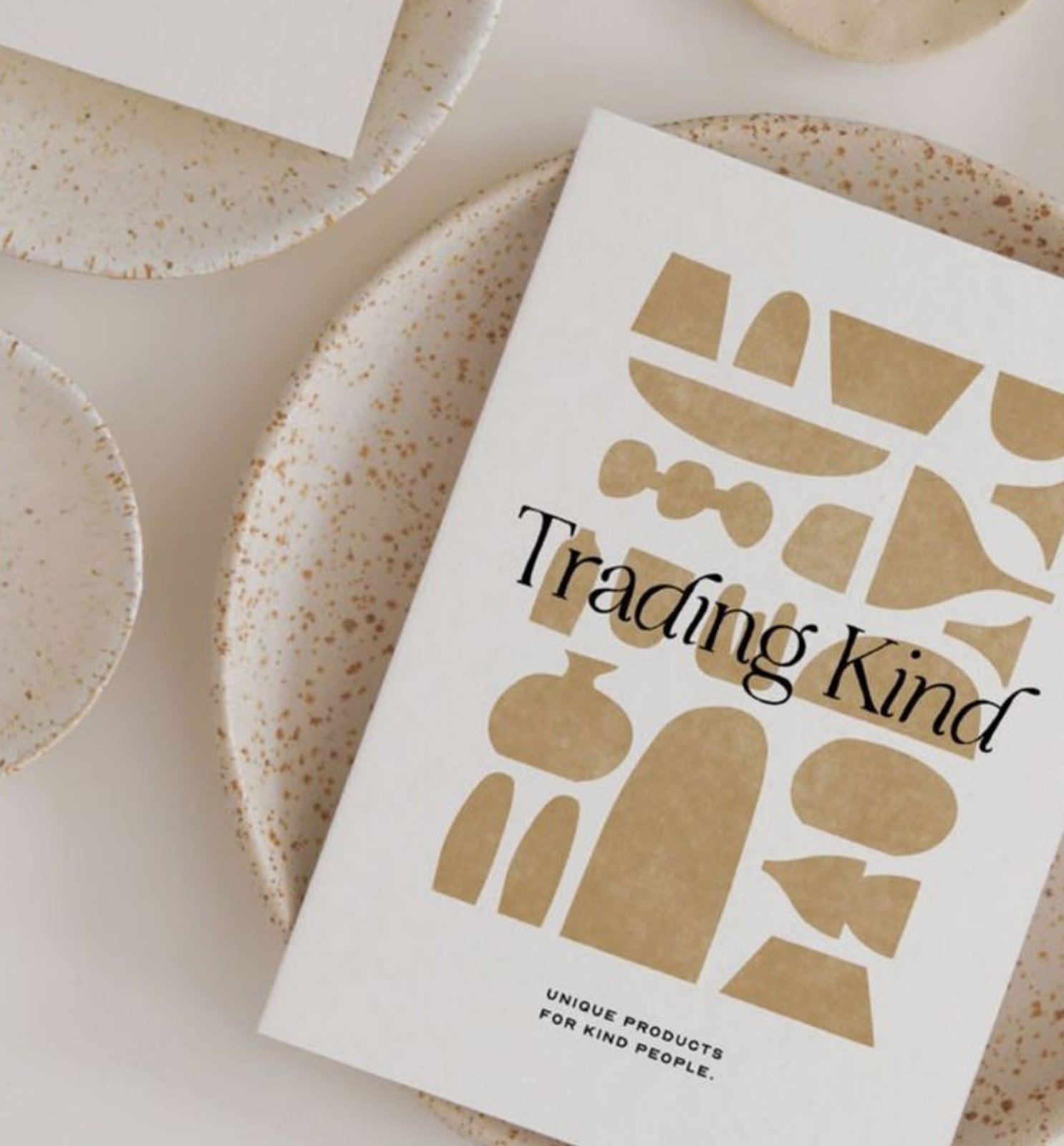 Close-up of a product packaging with abstract gold shapes and the text 'Trading Kind' against a white background, placed on speckled ceramic plates.