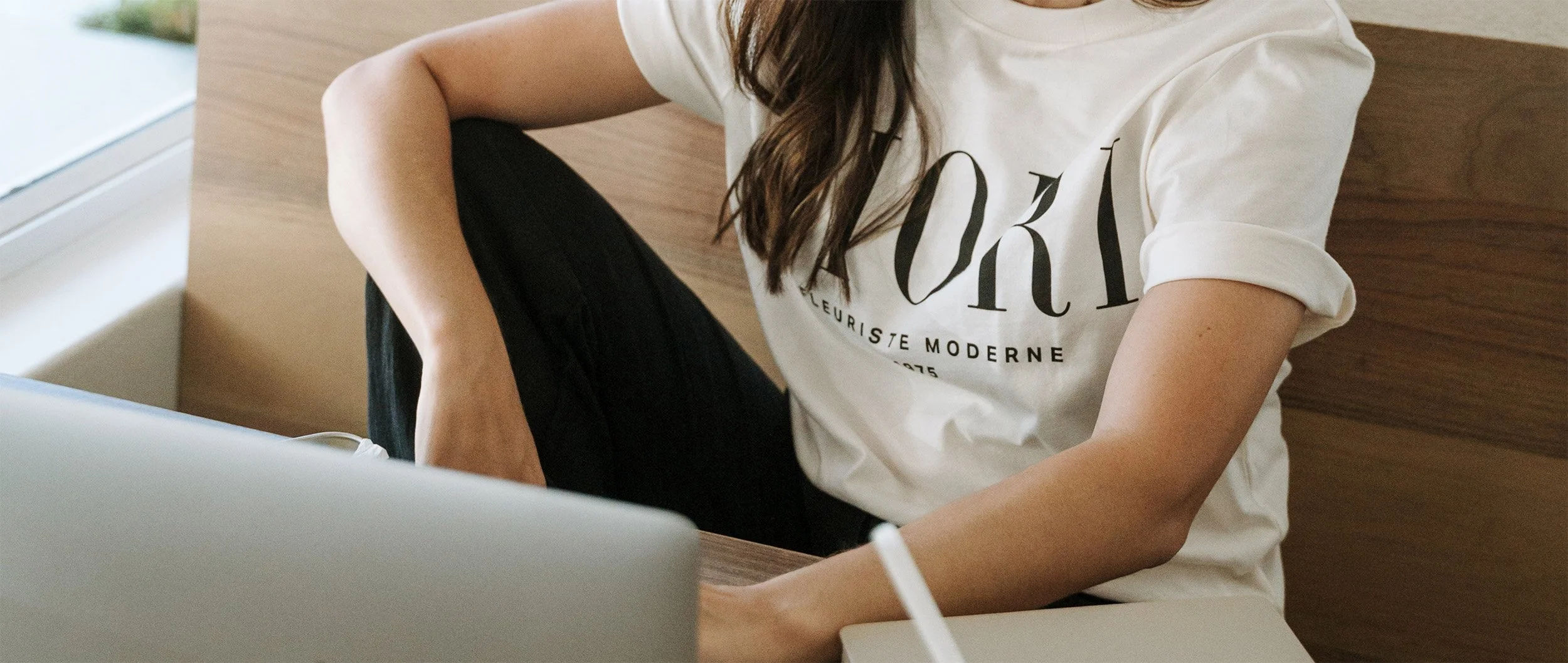 A person working on a laptop, wearing a white T-shirt with black text, sitting at a wooden desk in a well-lit room.
