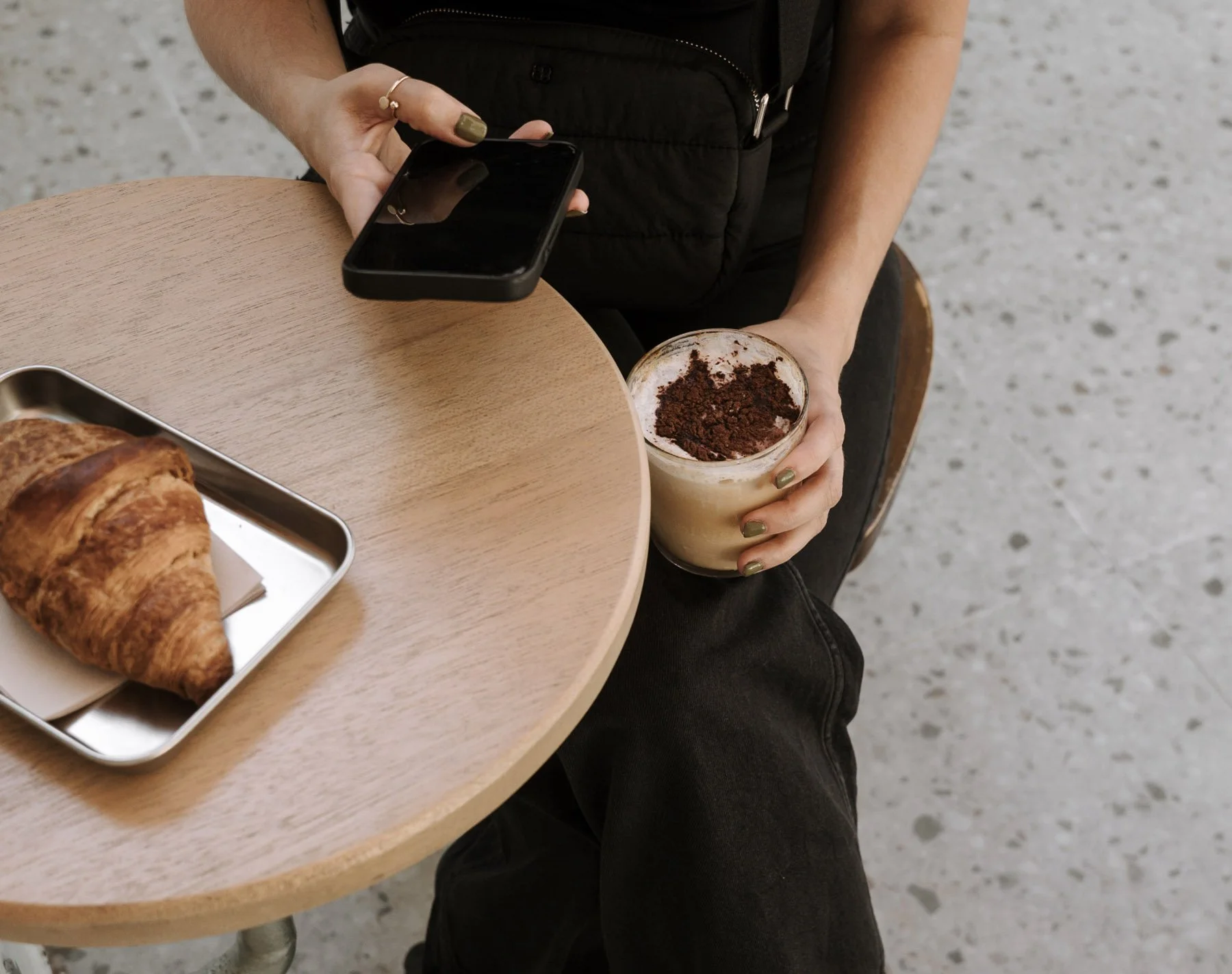 Person holding a smartphone in one hand and a glass of iced coffee topped with cocoa powder in the other hand, seated at a wooden table with a croissant on a tray.