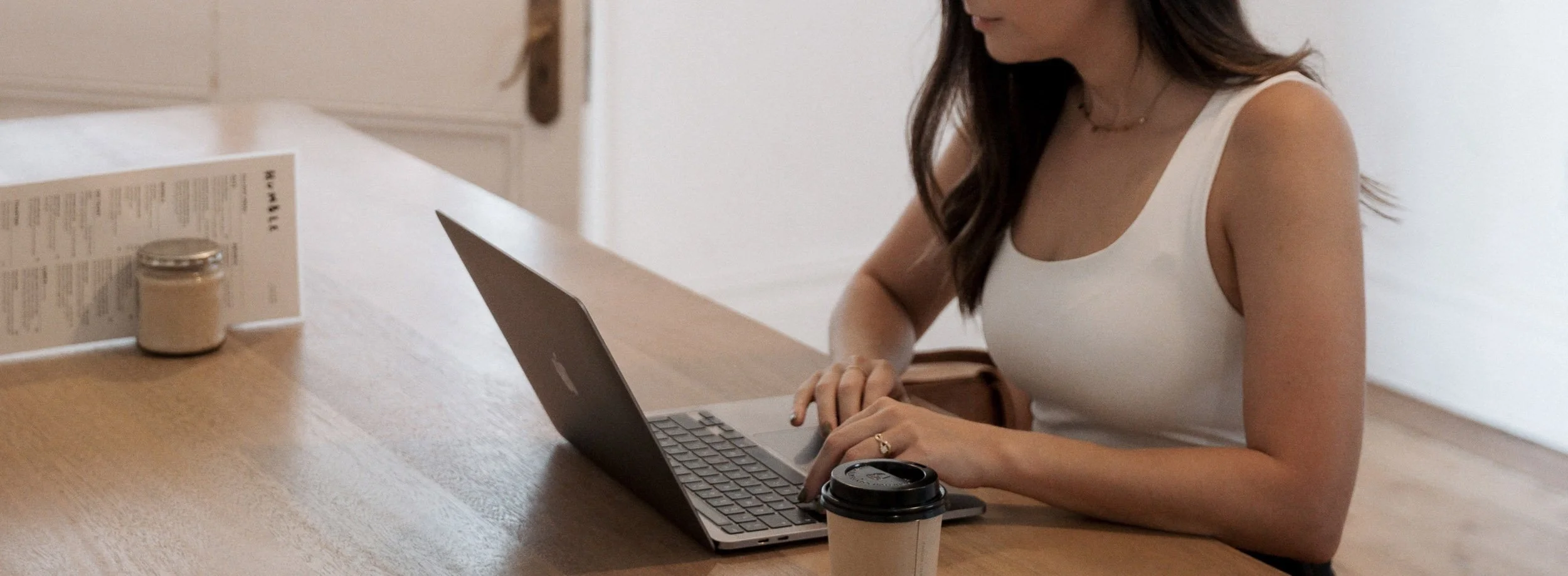 A woman in a white sleeveless top sitting at a wooden counter working on a laptop, with a paper coffee cup in front of her and a menu and jar to her left.