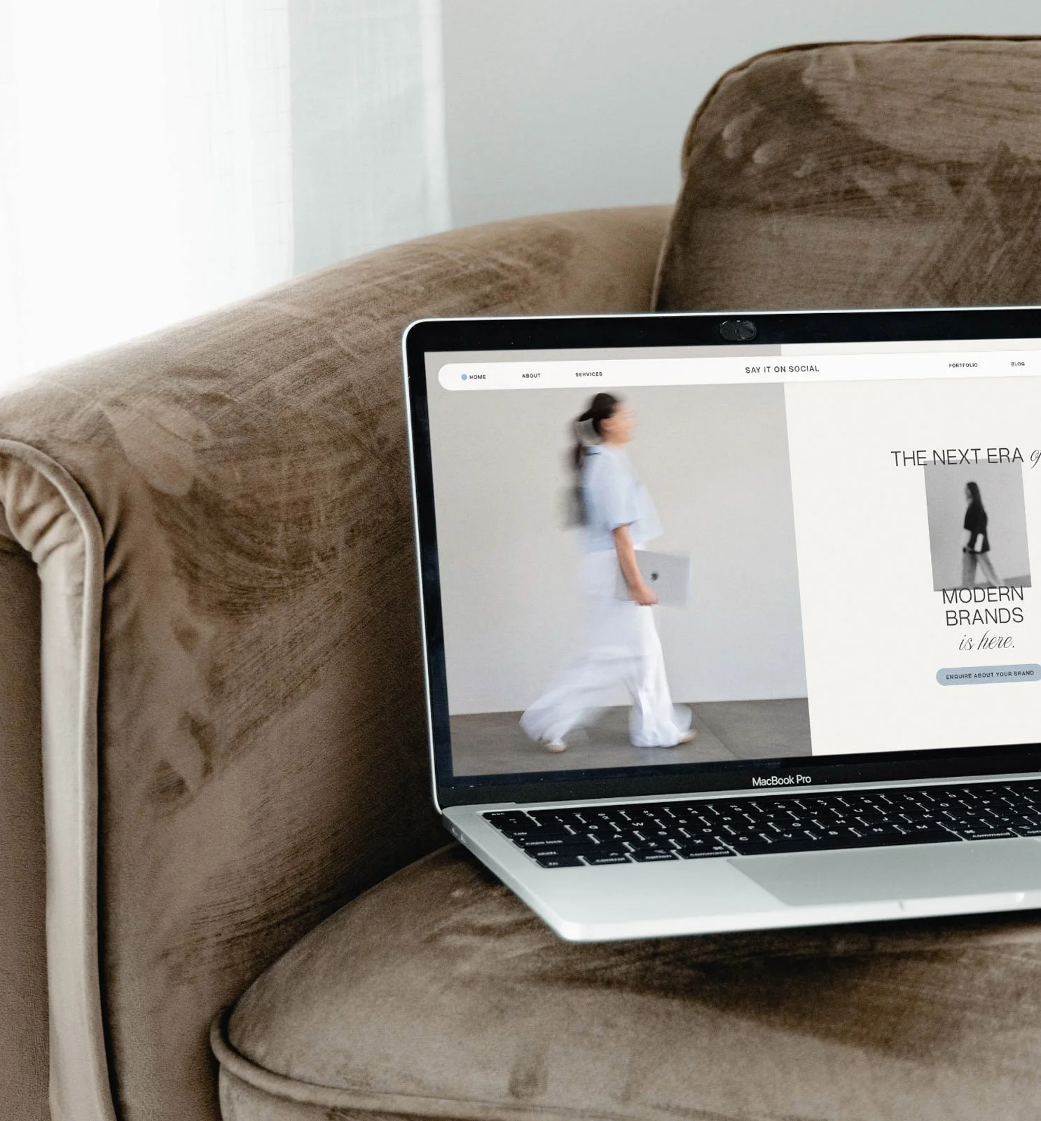 Partial view of a laptop on a beige sofa, showing a website with a woman in white clothing walking. The sofa has a velvety texture and there is a sheer white curtain in the background.