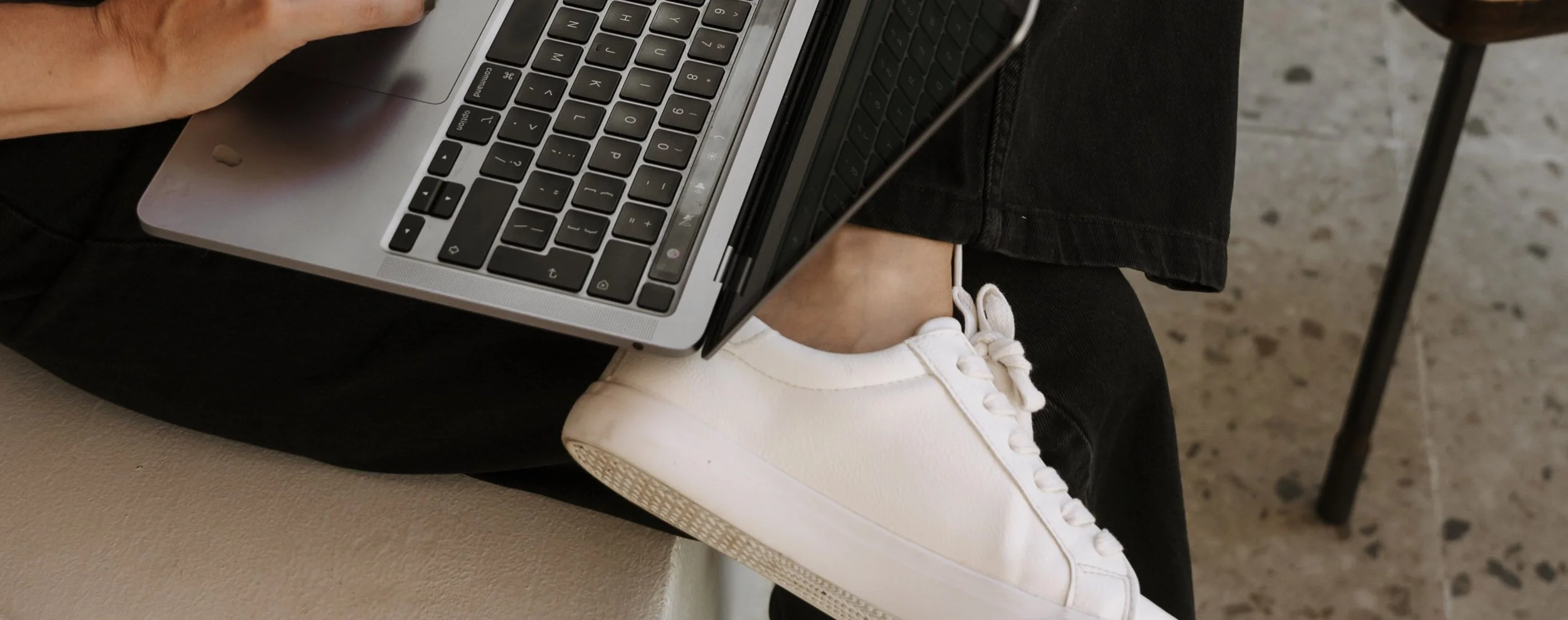 A person sitting with their legs crossed, wearing black pants and white sneakers, using a silver laptop on their lap. They are in an indoor setting with a speckled floor.