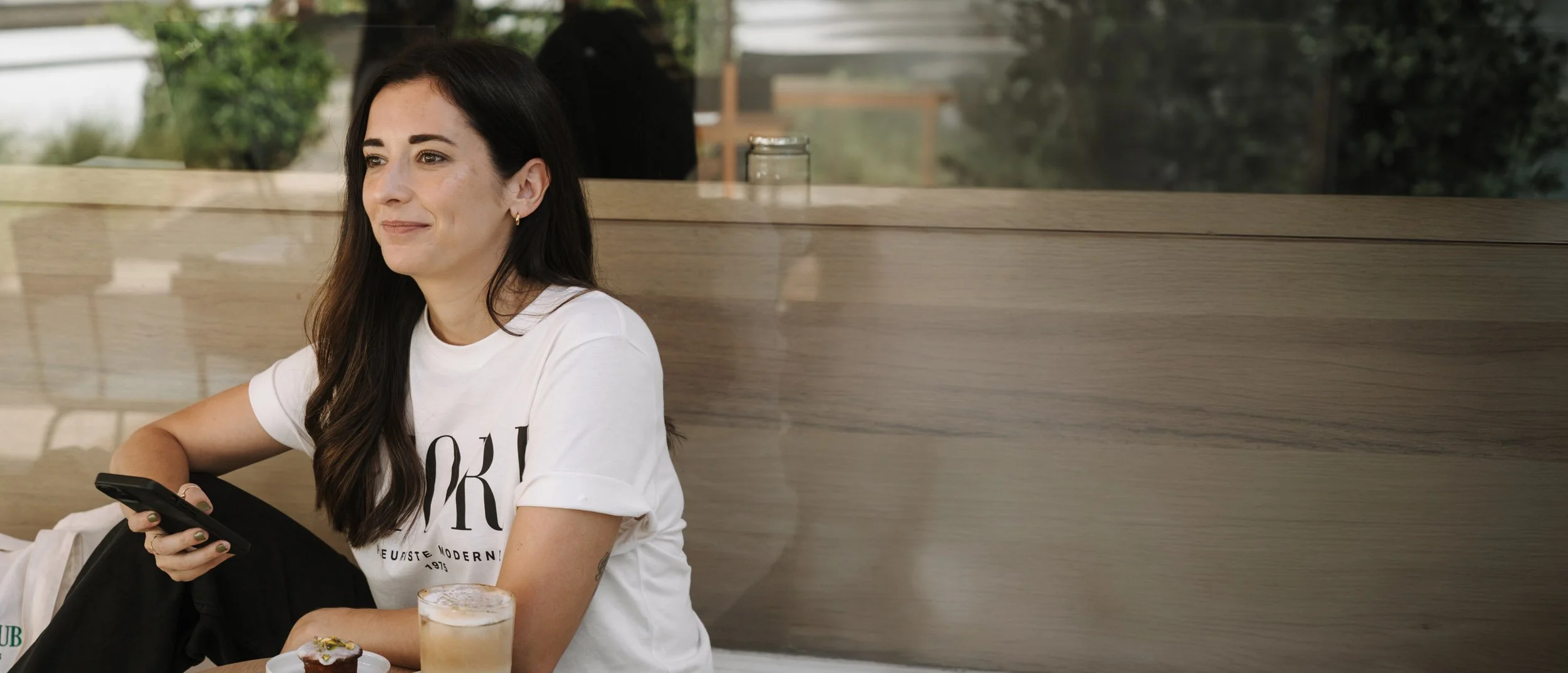 A woman with long dark hair and a white T-shirt sitting at a table in a cafe, holding a phone in her hand. There is a drink and a cupcake on the table. She is looking off to the side and smiling slightly.