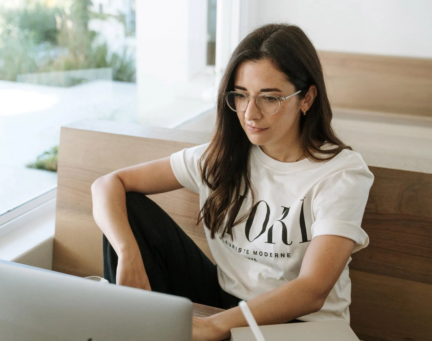 A woman with long dark hair, glasses, and earrings sitting at a ham, looking at a laptop with a window and greenery outside.